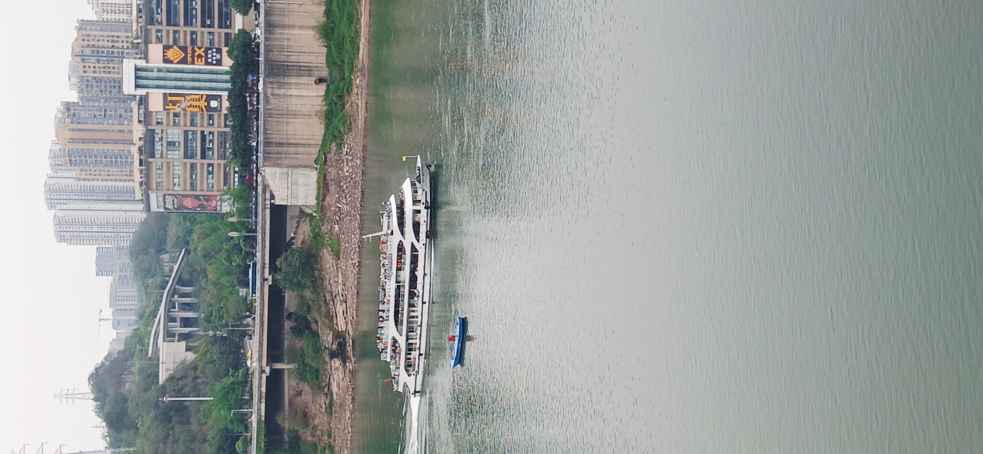 The image shows a boat on a river in Yuzhong District, China, with a city skyline on the left side of the river. The boat is a large, white passenger ferry filled with people, with a smaller blue boat trailing behind. The river is calm and the water is a gray-green color. On the left bank of the river, there is a mix of urban development, including high-rise buildings, roads, and green spaces. The sky is overcast and the lighting is diffused.