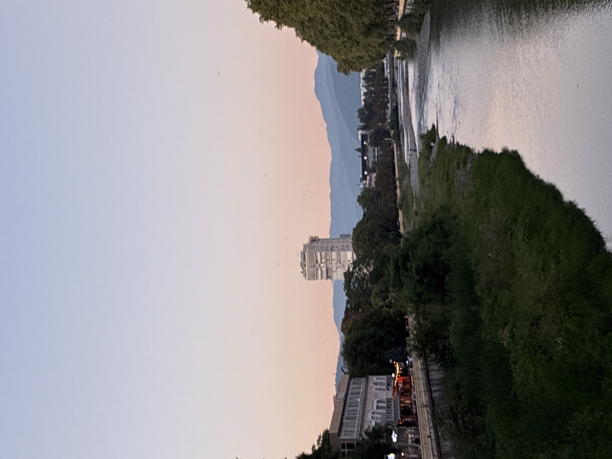 The image depicts a scenic view of the Mzimta River in Sochi, Russia, during the late evening. The sky is a gradient of pale blue and pink, with a few distant birds visible. On the left bank, a multi-story building with a modern facade stands next to older, more ornate structures. Lights are visible in the windows of the buildings, and a few people can be seen walking along the riverfront promenade. Lush green trees line the river, providing a natural contrast to the urban landscape. The Mzimta River flows through the scene, its surface reflecting the soft colors of the sky. In the background, a series of layered mountains fades into the distance, adding depth and grandeur to the composition. A tall, slender building, likely an apartment complex or hotel, is prominent in the mid-ground, with a distinctive architectural style. The overall impression is one of a serene and beautiful urban environment by the river.