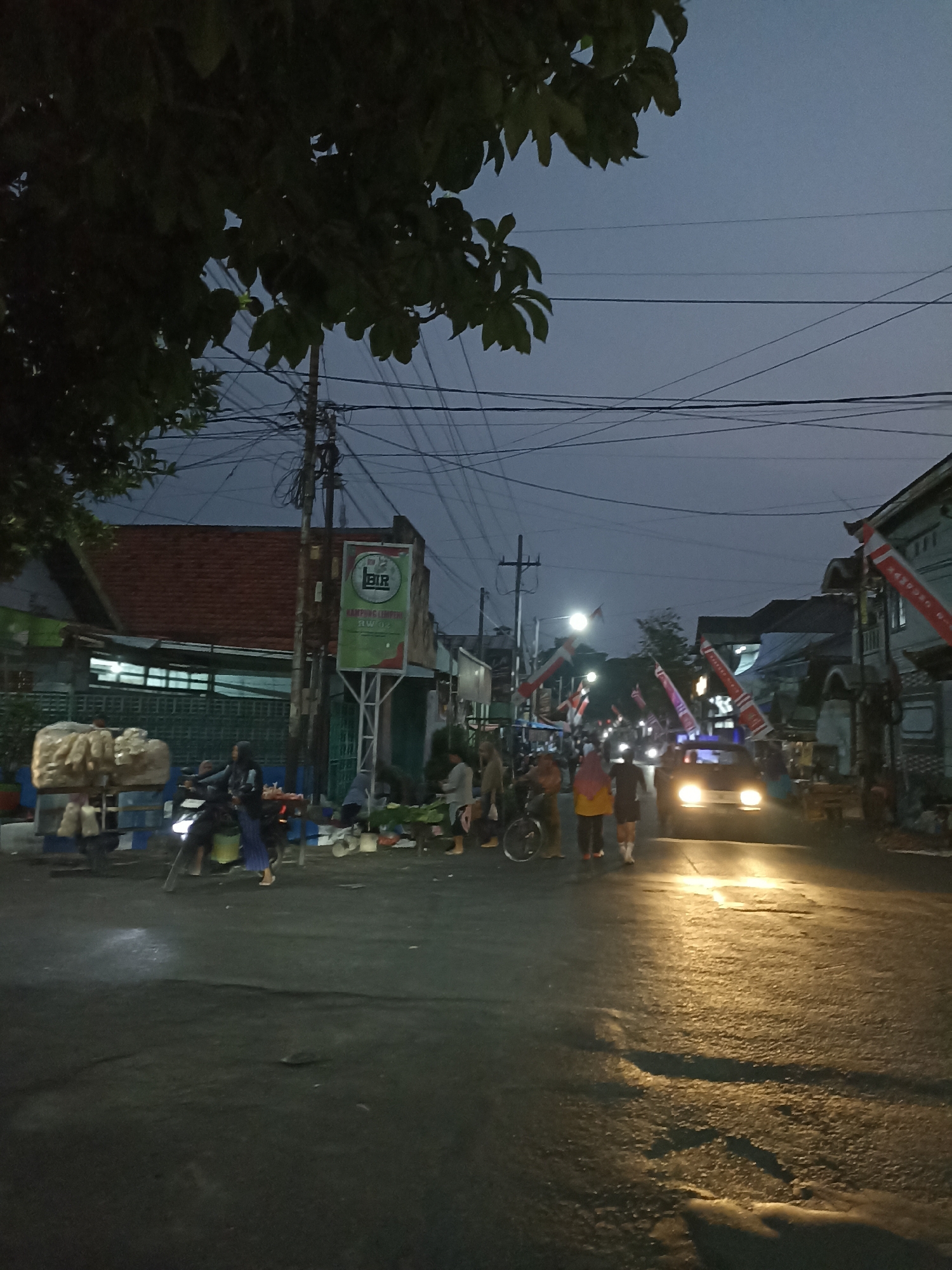 The image depicts a street scene in Ditotrunan, Indonesia, at dusk or early evening. The sky is a dusky blue, and overhead power lines are visible, crisscrossing the frame. A large tree branch with dark green leaves overhangs the left side of the image.
In the foreground, the asphalt road is wet and reflects the headlights of a vehicle and streetlights. Several people are present, some walking, others gathered in small groups. A motorcycle with two occupants is on the left side of the road. A car with its headlights on is moving down the street.
On the sides of the road, there are buildings and market stalls. One stall on the left displays a large pile of what appears to be food items, possibly snacks or produce, wrapped in plastic. A signpost with green and white branding is visible near the center of the image, reading "BIR" and "KAMPUNG CEMPLON". Red and white Indonesian flags on poles are attached to buildings and extend over the street in several locations, suggesting a local celebration or event. The overall atmosphere is that of a busy street at the transition from day to night, with community activity.