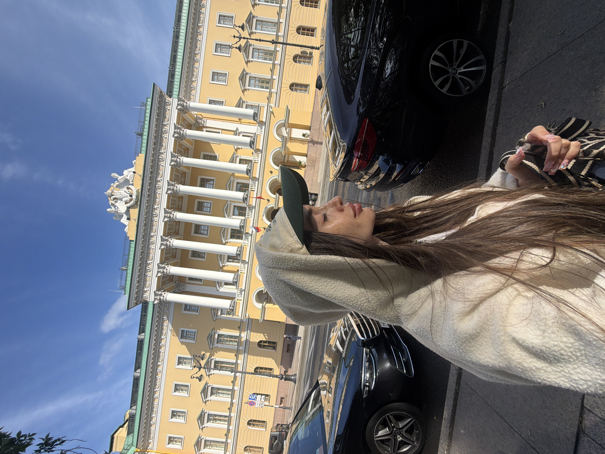 A young woman, wearing a light-colored hooded jacket and a baseball cap, is captured in a profile view, looking to the right. Her long, brown hair is visible. In the background, a grand, yellow building with classical architecture, featuring numerous columns and windows, stands under a bright blue sky with scattered white clouds. Several black cars are parked along the street in front of the building. The scene appears to be a street in Saint Petersburg, Russia, likely near a significant historical or governmental structure, given the building's facade. The lighting suggests it is daytime, with sunlight casting shadows.