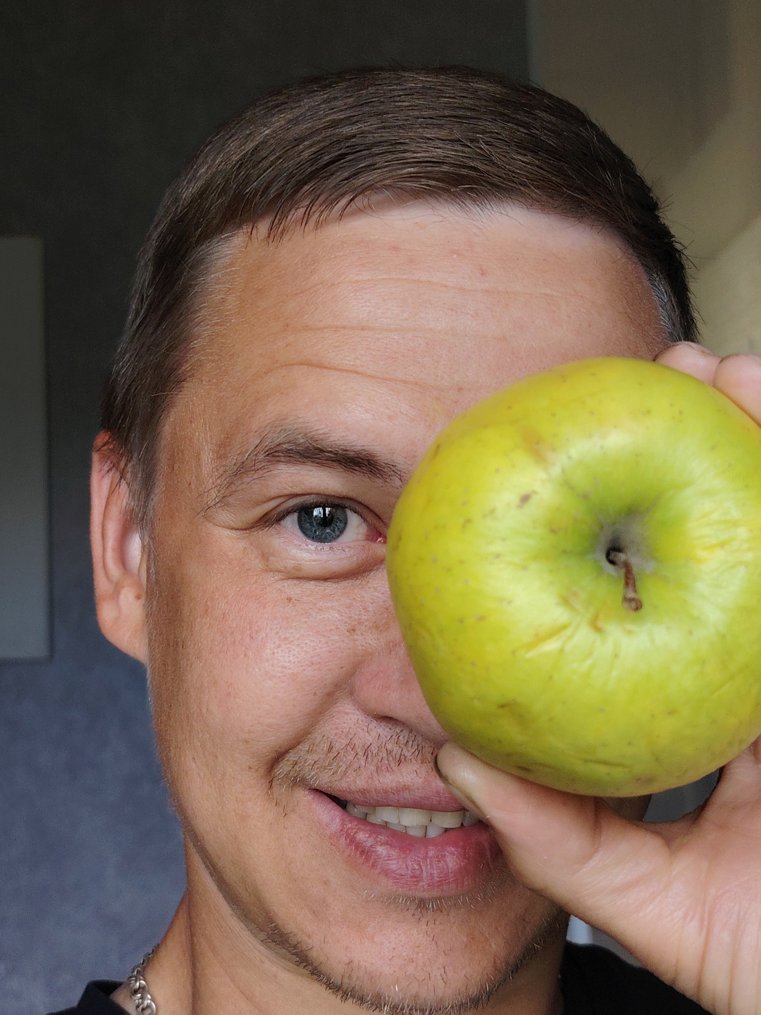In a close-up, waist-up shot, a person's face is partially obscured by a bright yellow apple held in their right hand. The person, with short brown hair and light skin, is looking towards the camera, with their left eye visible and blue in color. Their mouth is slightly open, revealing white teeth and pink lips. The background is a muted grey wall. The image has been taken in the city of Chaikovsky, Russia.