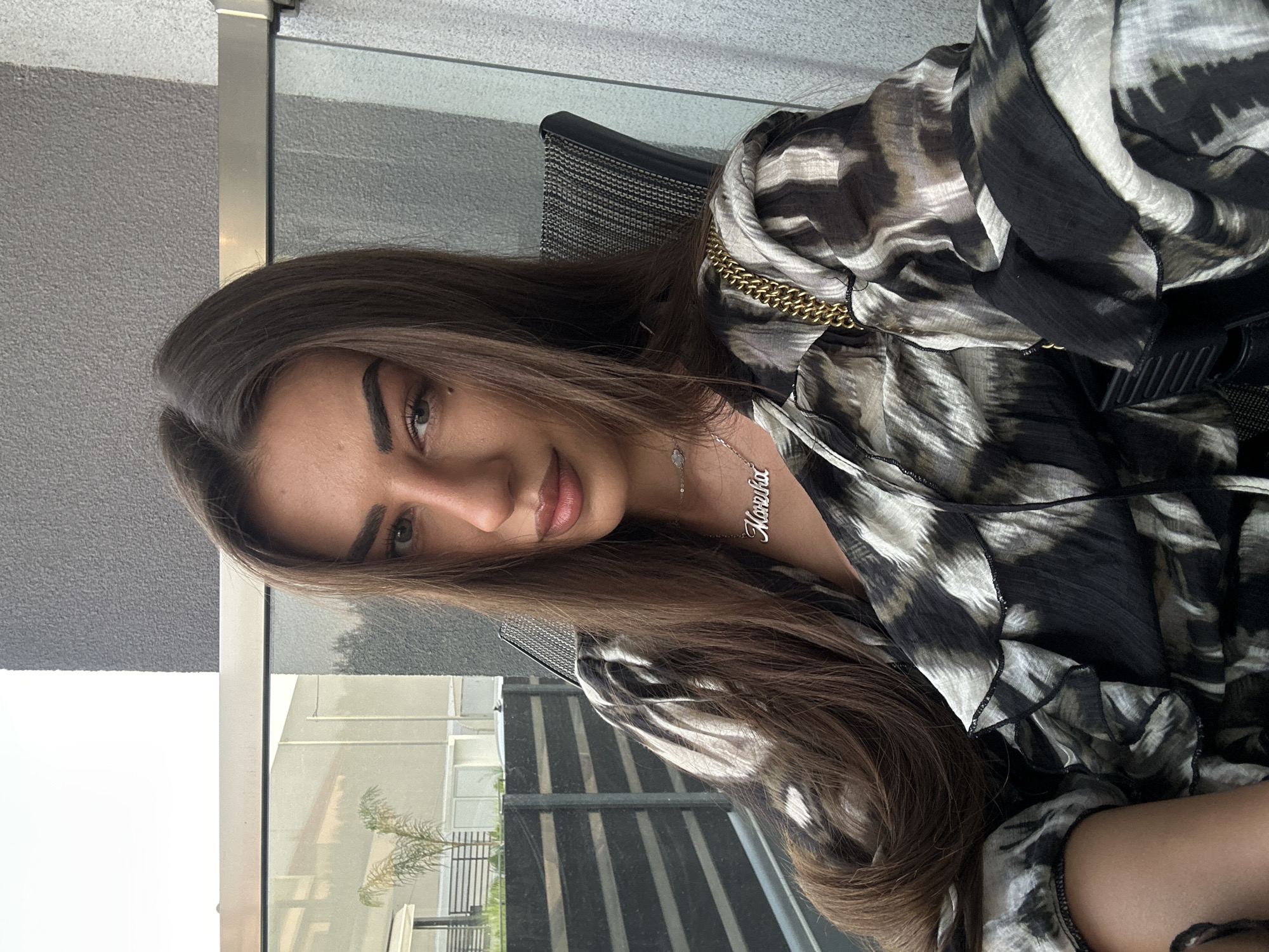 In Ofrynio Beach, Greece, a woman with long, dark brown hair sits in a chair on a balcony. She is wearing a black and white patterned blouse with ruffled sleeves and a gold chain purse slung over her shoulder. A silver necklace with the name "Mariha" hangs around her neck. The background shows a textured grey wall and a glass balcony railing, with a glimpse of an outdoor area featuring a small palm tree and a striped fence.