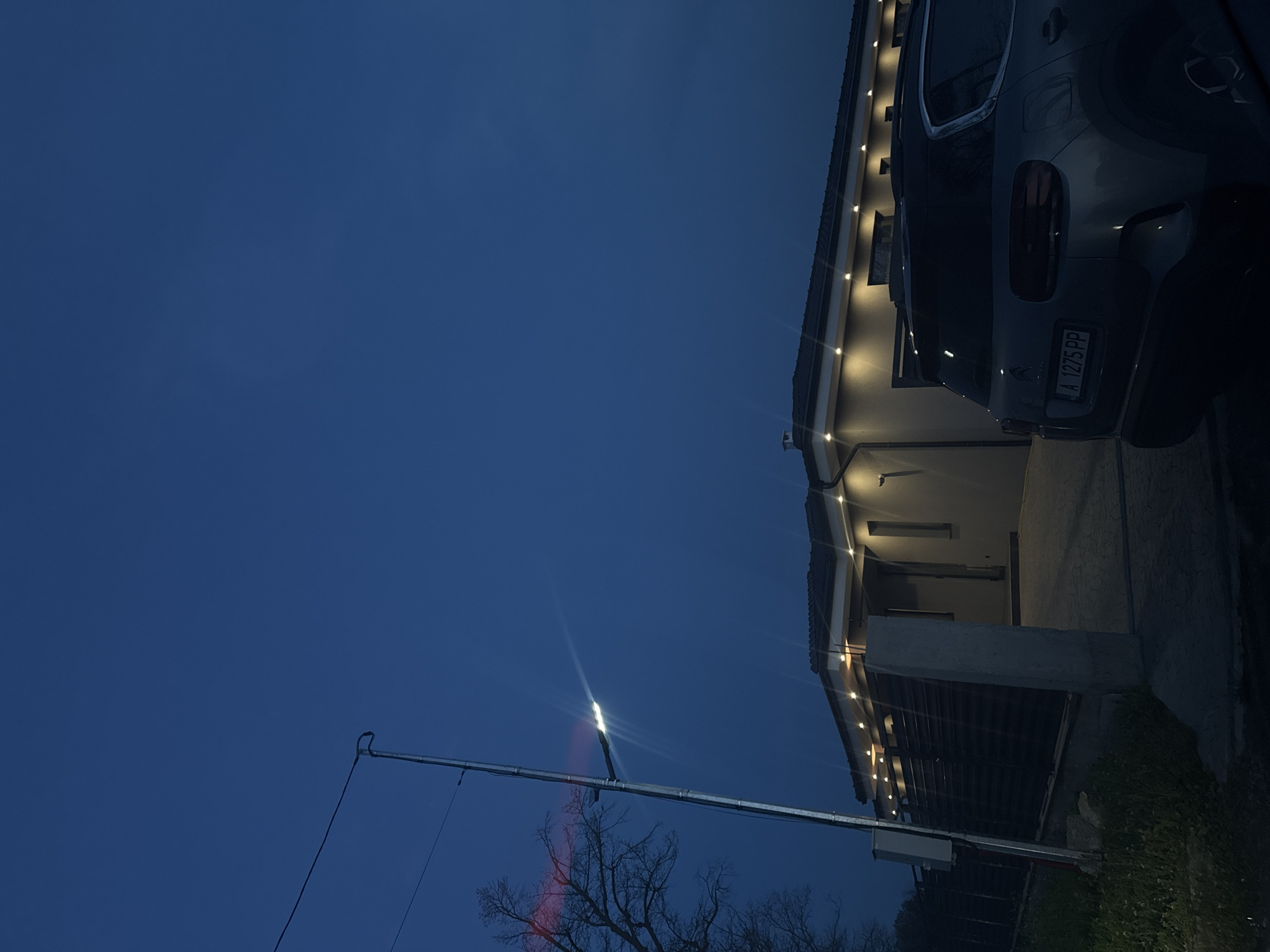 At dusk, the image shows a two-story residential building with outdoor lighting, possibly in Trastikovo, Bulgaria. The building is on the right side of the frame, and a car is parked near its entrance. A street light extends from the left of the frame towards the building. A leafless tree is visible beneath the street light. The sky is dark blue. The license plate on the car is A 1275 PP.