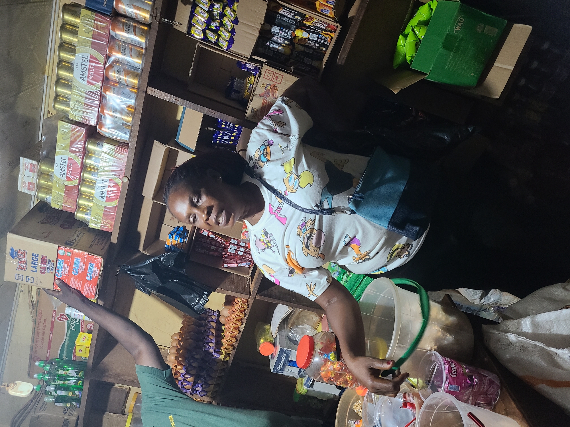 The image depicts an indoor scene, likely a small shop or market stall, with an abundance of products displayed on shelves and in boxes. The environment appears somewhat cluttered and dimly lit, with overhead lighting casting shadows.
In the foreground, a woman with dark skin and braided hair is looking upwards and to her right, with her mouth slightly open as if speaking. She is wearing a white t-shirt adorned with cartoon characters and a blue crossbody bag. Her left hand is extended and appears to be interacting with a green-rimmed bucket filled with goods. Next to this bucket, there are two clear plastic cups, one containing a pink item.
To the left of the woman, a person wearing a green t-shirt is partially visible. Their left arm is raised, holding a box labeled "YALE LARGE CABIN SWEETENED BISCUIT." Below this, stacked in trays, are numerous brown eggs.
The background is filled with shelves stocked with various items. Prominently displayed on the upper shelves are multiple packs of "AMSTEL MALTA" cans. Below these, there are boxes of what appear to be snacks or beverages, with some branded packaging visible, including "SOYA" and "XTREME." Several plastic bottles and containers are also scattered throughout the shelves. On the right side of the image, a green box with "WAO" branding is open, containing several green packets. A large, open black plastic bag hangs from one of the shelves.
The overall impression is of a busy retail space with a variety of food and drink items for sale. The presence of eggs, biscuits, canned beverages, and packaged snacks suggests a general provisions store. The visual elements do not provide specific clues to pinpoint the location to Abuja, Nigeria, beyond it being an indoor commercial setting.
