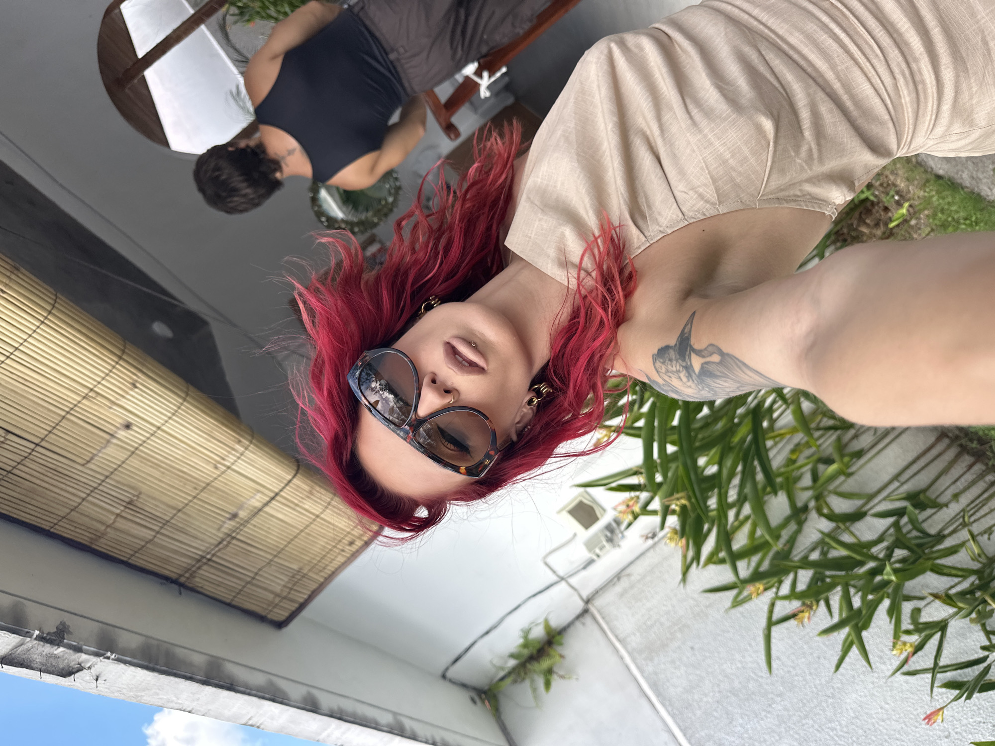 A photograph taken in North Kuta, Indonesia, features a woman with red hair, sunglasses, and multiple piercings, including a lip ring and earrings. She is wearing a light-colored, textured garment and has a tattoo on her upper arm. She appears to be holding the camera, possibly taking a selfie.
In the background, another individual wearing a dark tank top and brown shorts is seated, and there is a white-topped table with what appears to be a wooden frame. The environment includes a bamboo-style window covering, a white building exterior, and greenery, suggesting an outdoor or semi-outdoor setting. The perspective is looking upwards. The sky is visible in the bottom left corner.