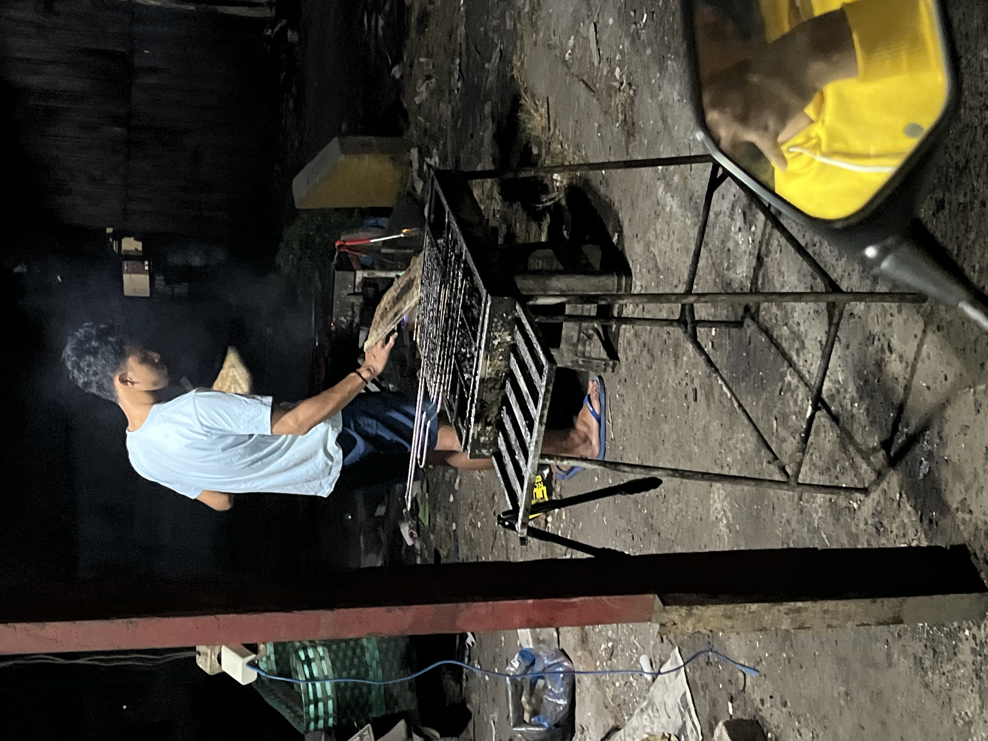 A man is grilling food outdoors at night, in the city of Kalinyamatan, Indonesia. He is wearing a light blue t-shirt and dark blue shorts, and flip-flops. He is holding a flat, wooden object, possibly a tray, over the grill, and appears to be tending to the food. There is smoke rising from the grill, indicating that the food is cooking. The grill is a metal structure with a grate, and it is placed on a stand. The ground around the grill is dirt and debris. In the foreground, a motorcycle mirror reflects the scene, showing a person in a yellow jacket. The environment appears to be somewhat unkempt, with various items scattered around. The lighting suggests it is nighttime, and the overall atmosphere is casual.