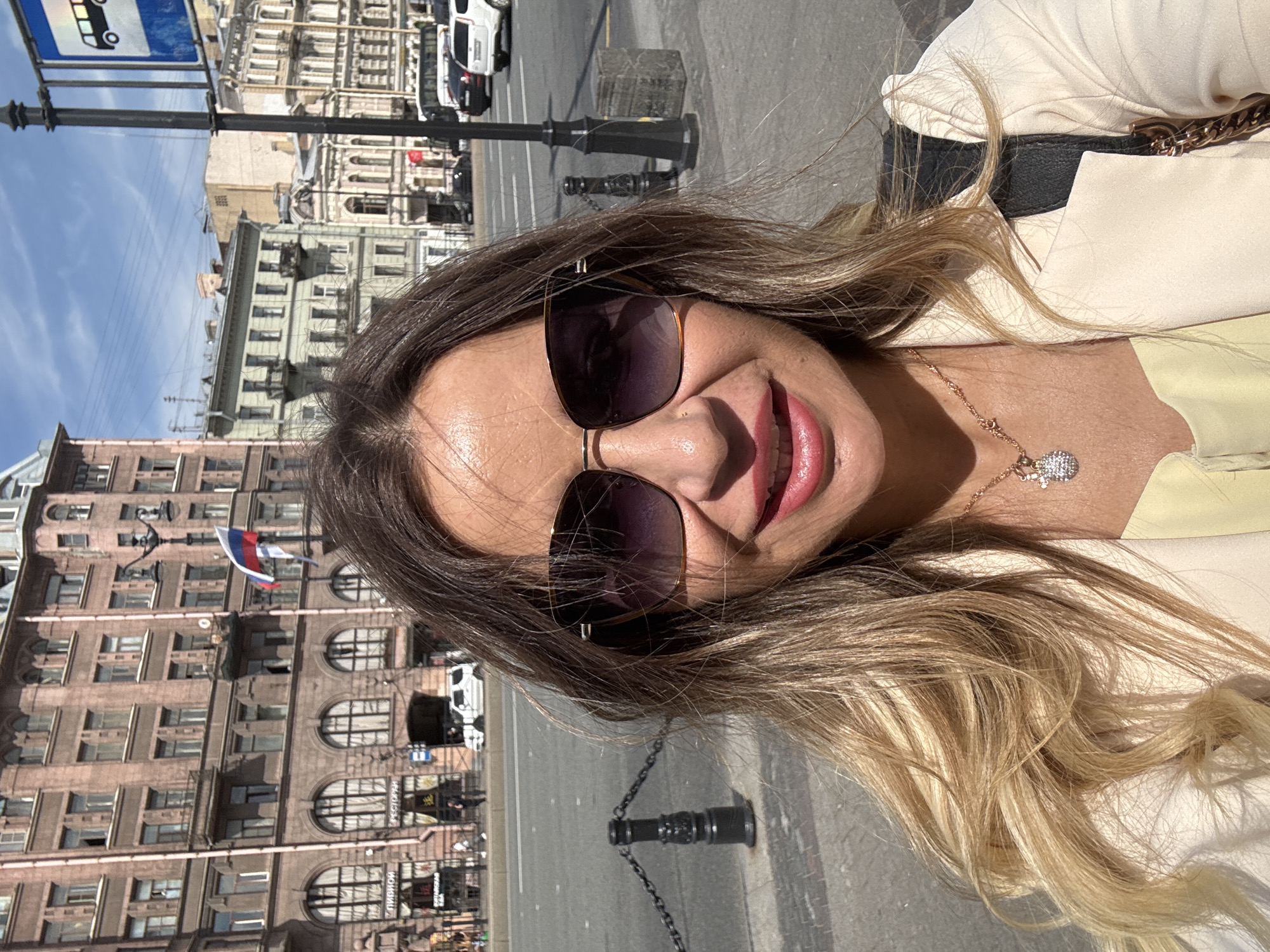 A woman is taking a selfie on a sunny day in Saint Petersburg, Russia. The background shows a street with several buildings and a flag. The woman is wearing sunglasses and a light-colored jacket over a yellow top. She has blonde hair and is wearing a necklace. In the background, there are cars, a bus stop sign, and a lamppost. The buildings are multi-story structures with arched windows and balconies. The architecture suggests an older, European style.