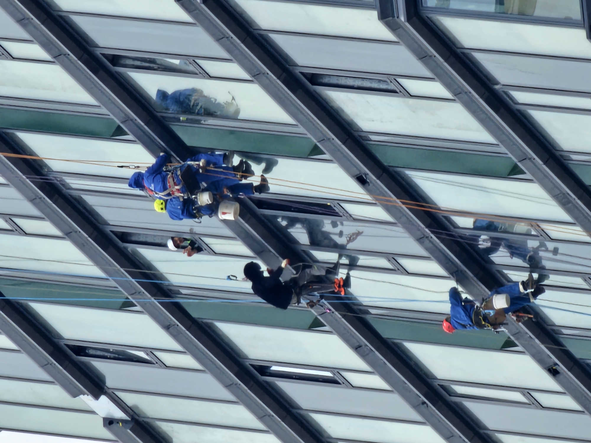 Four window washers are cleaning the exterior of a modern, glass-walled building. The building's facade features a grid-like pattern of windows separated by dark, angled beams.
The window washers are suspended by ropes and harnesses. One wears a yellow hard hat and blue uniform. Another wears a black outfit. The third wears a red hard hat and blue uniform. The fourth is wearing a blue uniform with a hood.
Reflections of the surrounding environment and sky are visible in the glass windows.