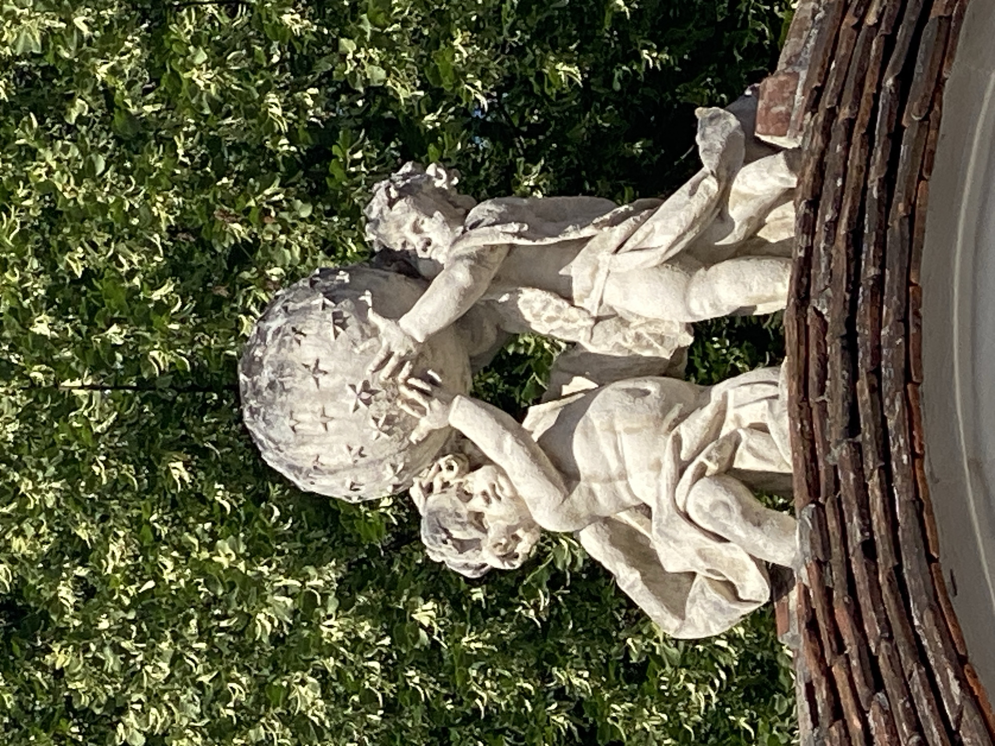 This is a photograph of a stone sculpture of two cherubic figures interacting with a globe. The figures are draped in flowing robes and appear to be straining to hold or manipulate the globe, which is decorated with star-like patterns. The sculpture is positioned outdoors, with lush green foliage serving as the backdrop. A portion of a textured, possibly wooden or tiled, structure is visible on the right side of the image, adding a rustic element to the scene. The lighting suggests it is daytime, with sunlight casting shadows and highlighting the details of the sculpture and the surrounding leaves. The overall impression is one of artistic endeavor and classical representation within a garden setting. There is no visible text in the image.