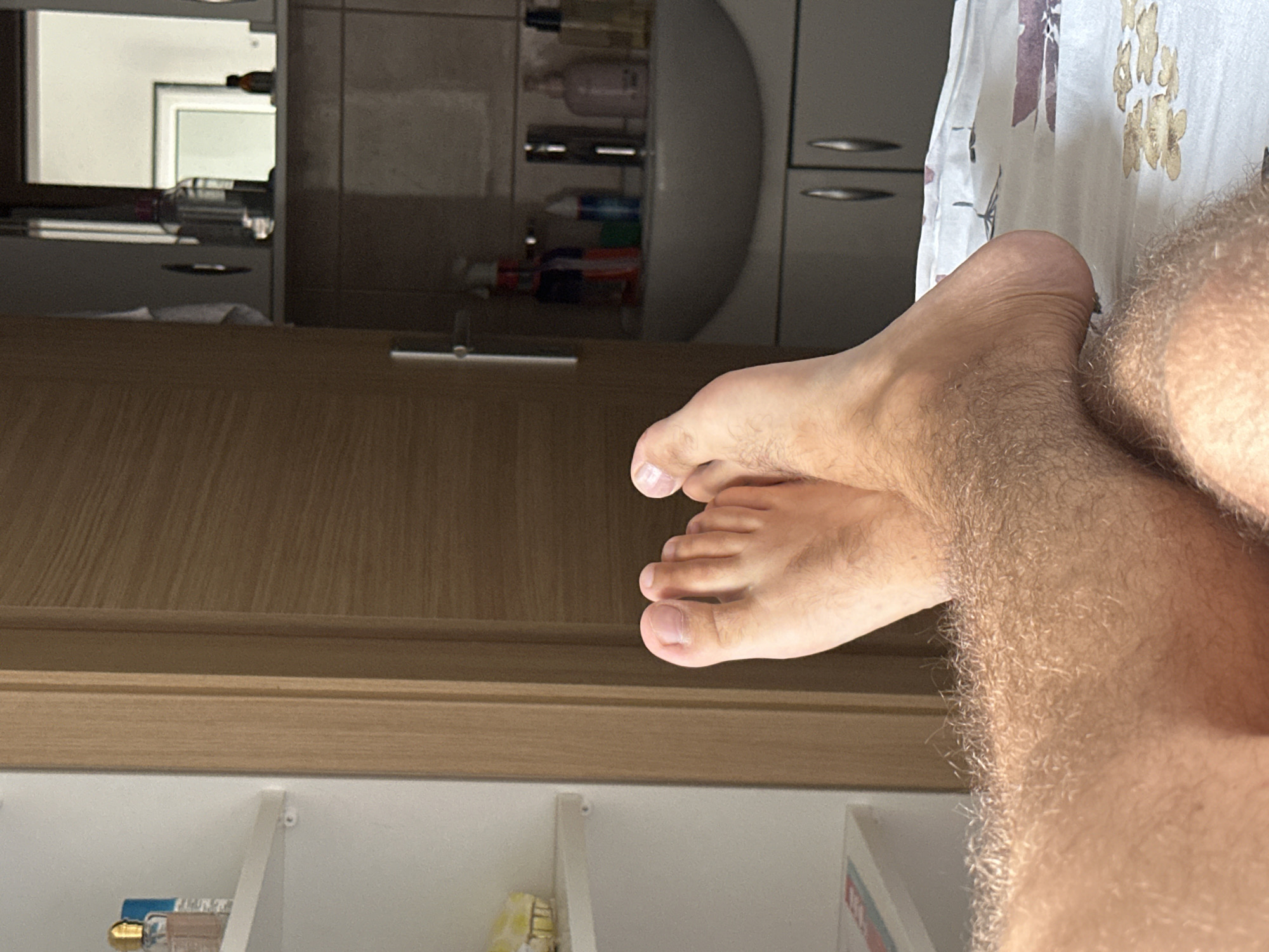 The image displays a close-up view of a person's bare feet resting on a bed with a floral patterned duvet. In the background, a wooden door is visible, and to the right, a bathroom area with a sink and various toiletries on a shelf. The overall setting suggests a domestic interior, possibly a bedroom in Budva, Montenegro. The lighting indicates daytime.