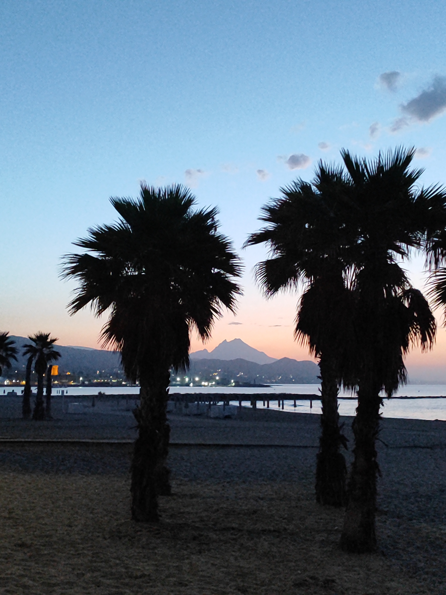 The image depicts a silhouetted scene of palm trees on a beach in El Campello, Spain, during dusk. The sky displays a gradient of blues and oranges, with a few scattered clouds. In the distance, mountains are visible, and a coastline with lights suggests a town or city. The foreground is a sandy beach with several palm trees casting long shadows. The ocean is calm and reflects the sky's colors. The overall atmosphere is peaceful and serene, capturing a moment of transition between day and night.