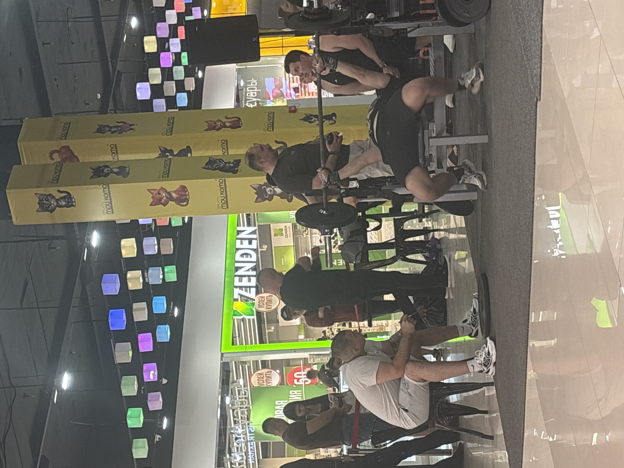 In a brightly lit indoor space, possibly a shopping mall or recreational facility in Tri Protoka, Russia, several individuals are engaged in various activities. In the foreground, a man in a light-colored t-shirt and shorts sits on a chair, holding a smartphone. Adjacent to him, another man is performing a bench press with a barbell. He is lying on his back on a bench, holding the barbell with both hands. A man in a dark t-shirt stands behind him, seemingly spotting or assisting. Further in the background, a group of people are visible, some standing and interacting, others appearing to be observing. The environment features distinctive cylindrical pillars decorated with cartoon cat imagery and the text "mou koma" in multiple languages. Overhead, colorful cube-shaped lights hang from the ceiling. The floor is a reflective surface, possibly polished concrete or tile, and a dark gray mat is laid out for the weightlifting activity. A green sign with the word "ZENDEN" is prominently displayed in the background.