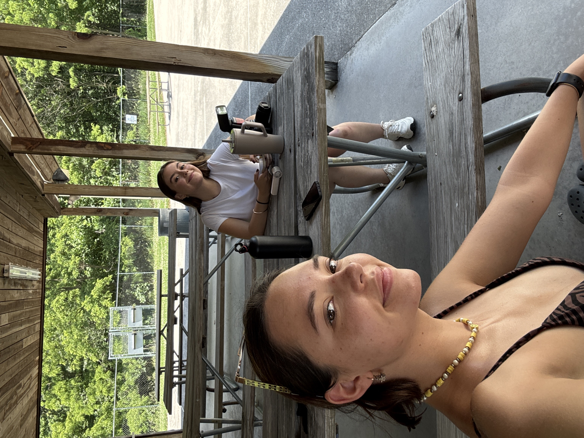 In Washington Township, two young women are seated at a wooden picnic table under a covered pavilion. The woman in the foreground is looking at the camera and smiling, wearing a brown and black striped top and a yellow beaded necklace. She has sunglasses perched on her head and a star-shaped earring in her left ear. The woman in the background is also looking at the camera and smiling, wearing a white t-shirt and white sneakers. On the table between them are a large gray travel mug, a camera with a flash attachment, and a black water bottle. The pavilion has a wooden roof and wooden support beams. Beyond the pavilion, a chain-link fence encloses a grassy area with trees.