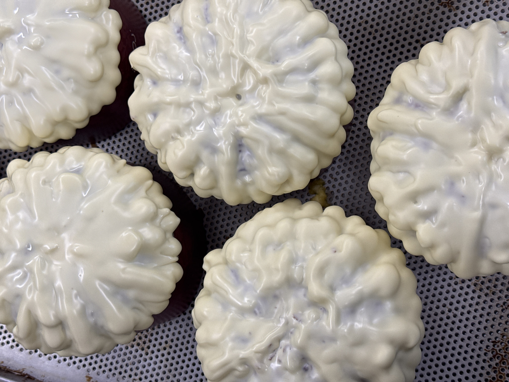 The image shows five circular pastries covered in a glossy white frosting. The pastries are arranged on a perforated metal tray. The pastries have a slightly ruffled or textured appearance due to the frosting application. The background is indistinct, suggesting the focus is on the pastries themselves. The image does not contain enough information to determine a specific location in Belgrade, Serbia, or to identify any people, actions, or interactions.
