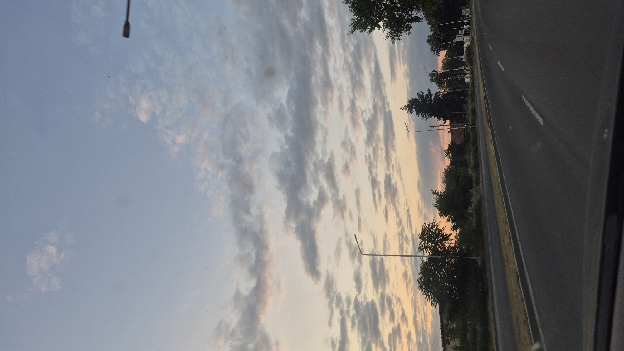 The image depicts a view from a moving vehicle on a road, likely an exit from or entrance to a city. The sky is filled with scattered clouds, tinged with the warm colors of a sunset or sunrise, transitioning from orange and pink near the horizon to a pale blue overhead. Streetlights are visible along the side of the road. Trees and some buildings are present on the left side of the road, indicating an urban or semi-urban setting. The road itself appears to be asphalt, with lane markings visible. The perspective suggests the photograph was taken from the driver's or front passenger's seat of a car. Given the context of Lovech, Bulgaria, this scene could represent a road leading into or out of the city, with the characteristic architecture and vegetation of the region.