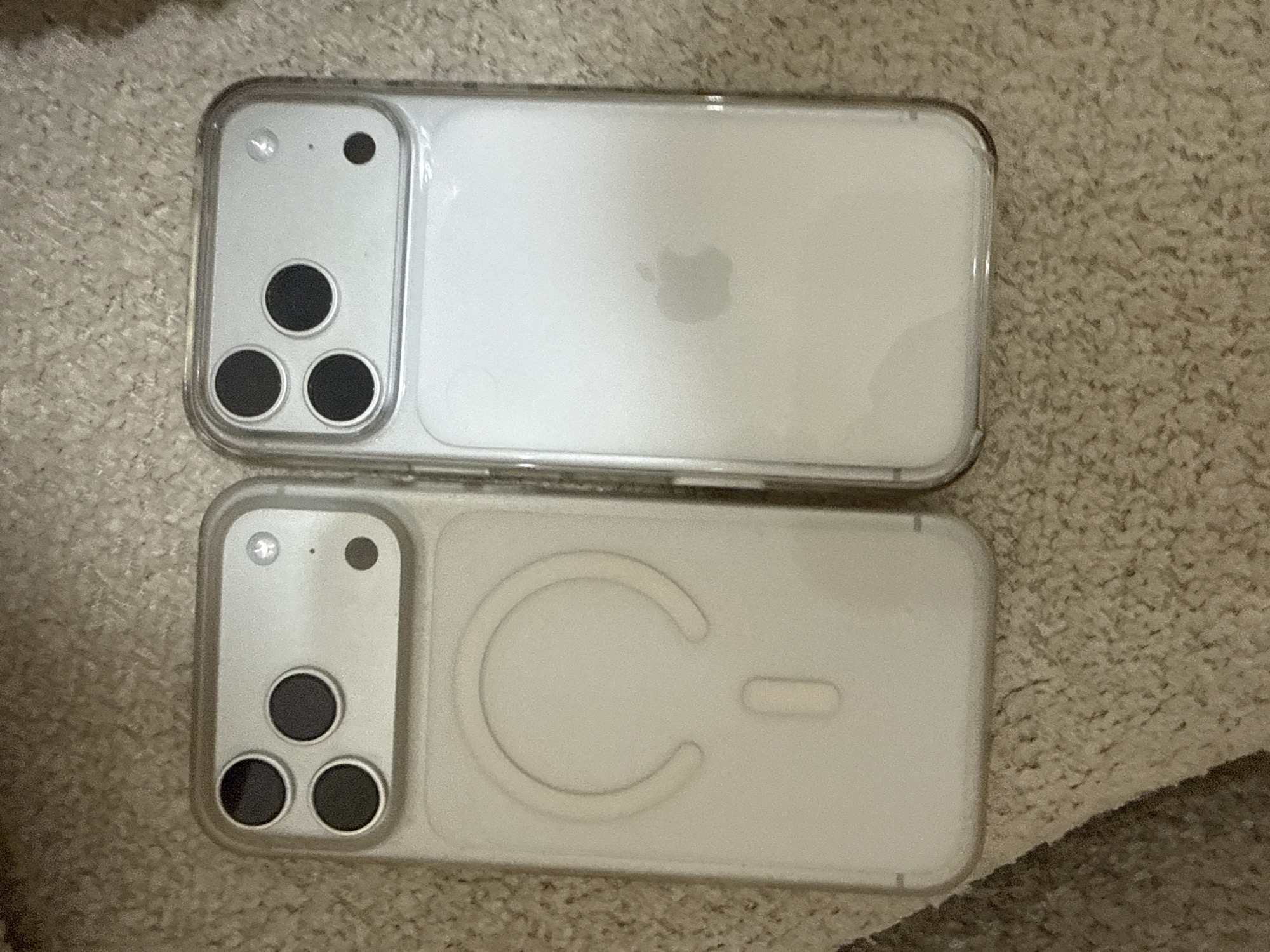The image shows two clear phone cases, each designed for an iPhone with a triple-lens camera system. The case on the left has a circular indentation and a smaller oval shape on its back, indicating it is compatible with MagSafe technology. The case on the right has a visible Apple logo on its back. Both cases are placed on a textured, carpeted surface that appears to be a floor. There are no people or any identifiable urban features in the image. The specific location in Tbilisi, Georgia, cannot be determined from the visual information provided.