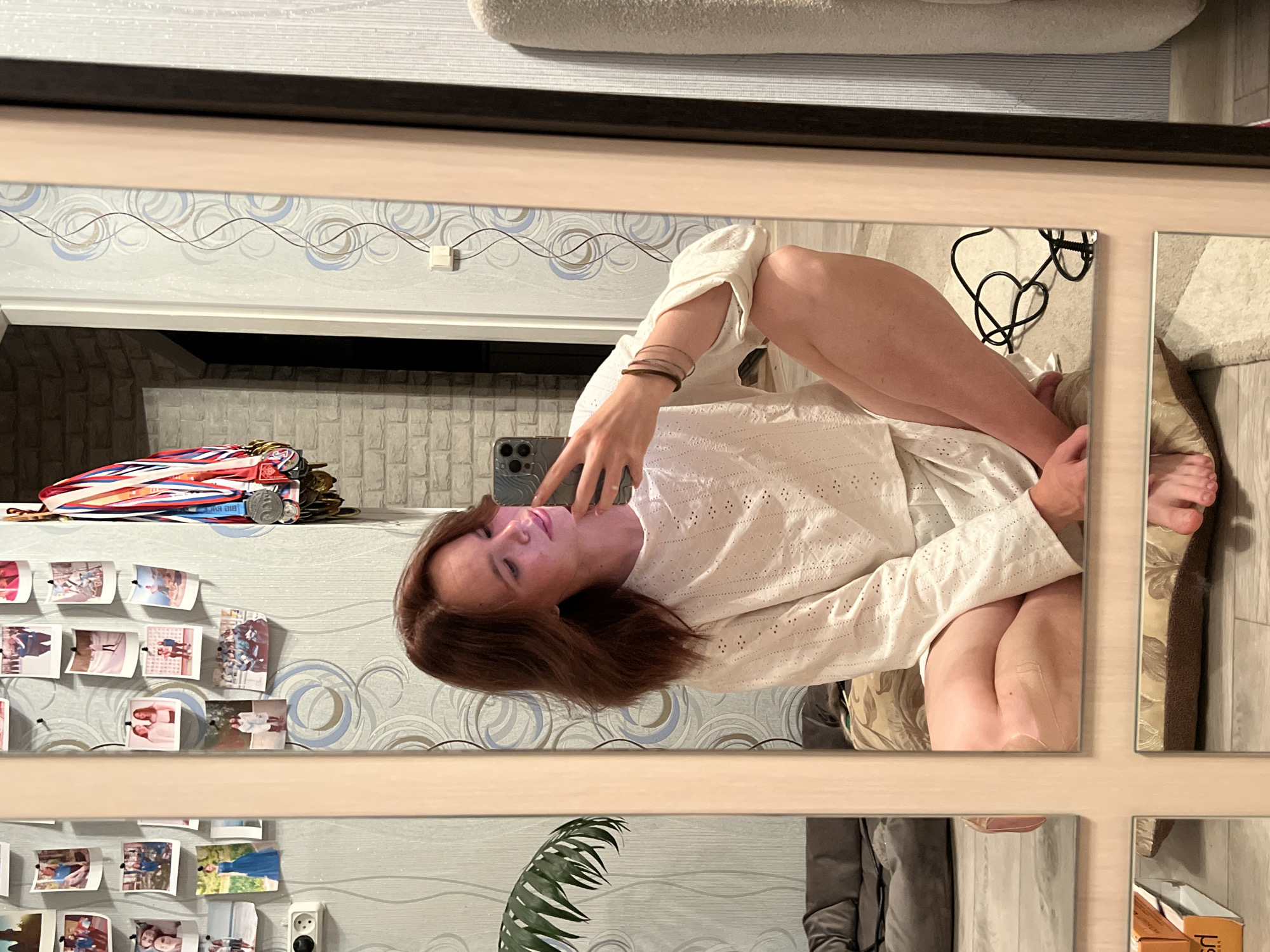 A woman is reflected in a mirror with multiple panes. She is wearing a white shirt and has brown hair. The room appears to be a bedroom. There are pictures and medals displayed on the wall. A plant is visible in one of the mirror panes. The location is Chaikovsky, Russia.