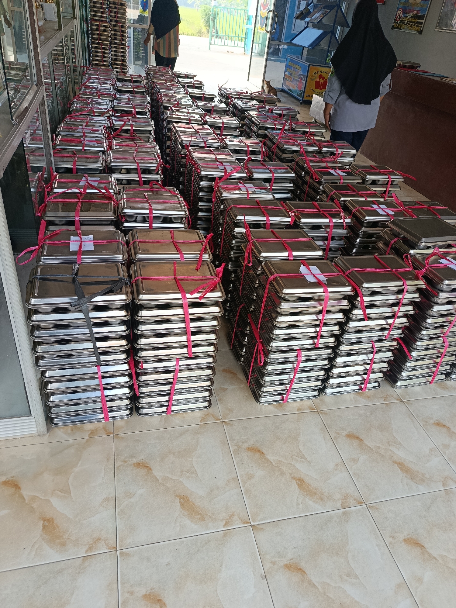 This image depicts an outdoor scene in Palembang, Indonesia, where stacks of metallic trays are neatly arranged on a tiled floor. The trays, uniformly rectangular, are bundled together with vibrant pink plastic straps. Some stacks are taller than others, creating a sense of abundance. In the background, two women are visible. One, wearing a black hijab and a grey uniform, stands near a doorway. Another woman, dressed in a striped top and headscarf, is also present. Behind them, a building with signage and a glimpse of greenery suggests an urban or semi-urban environment. The overall impression is one of organized preparation or distribution, with a focus on the numerous trays.