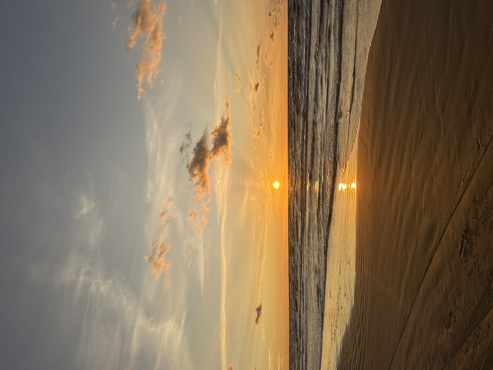 This is a vertical, wide-angle, eye-level shot of a beach at sunset, likely located in Jūrmala, Latvia, given its characteristic shoreline and the typical sunset colors observed over the Baltic Sea. The sky is a gradient of colors, transitioning from a soft grey at the top to a vibrant orange and yellow near the horizon. Wispy clouds are scattered across the sky, tinged with pink and orange from the setting sun. The sun itself is a bright, round orb low on the horizon, casting a shimmering golden reflection on the water. Gentle waves roll onto the shore, creating a foamy white line where they meet the sand. The beach is composed of fine, damp sand, with subtle ripples and tire tracks visible, indicating recent activity, though no people or animals are present. The overall scene evokes a sense of tranquility and natural beauty.