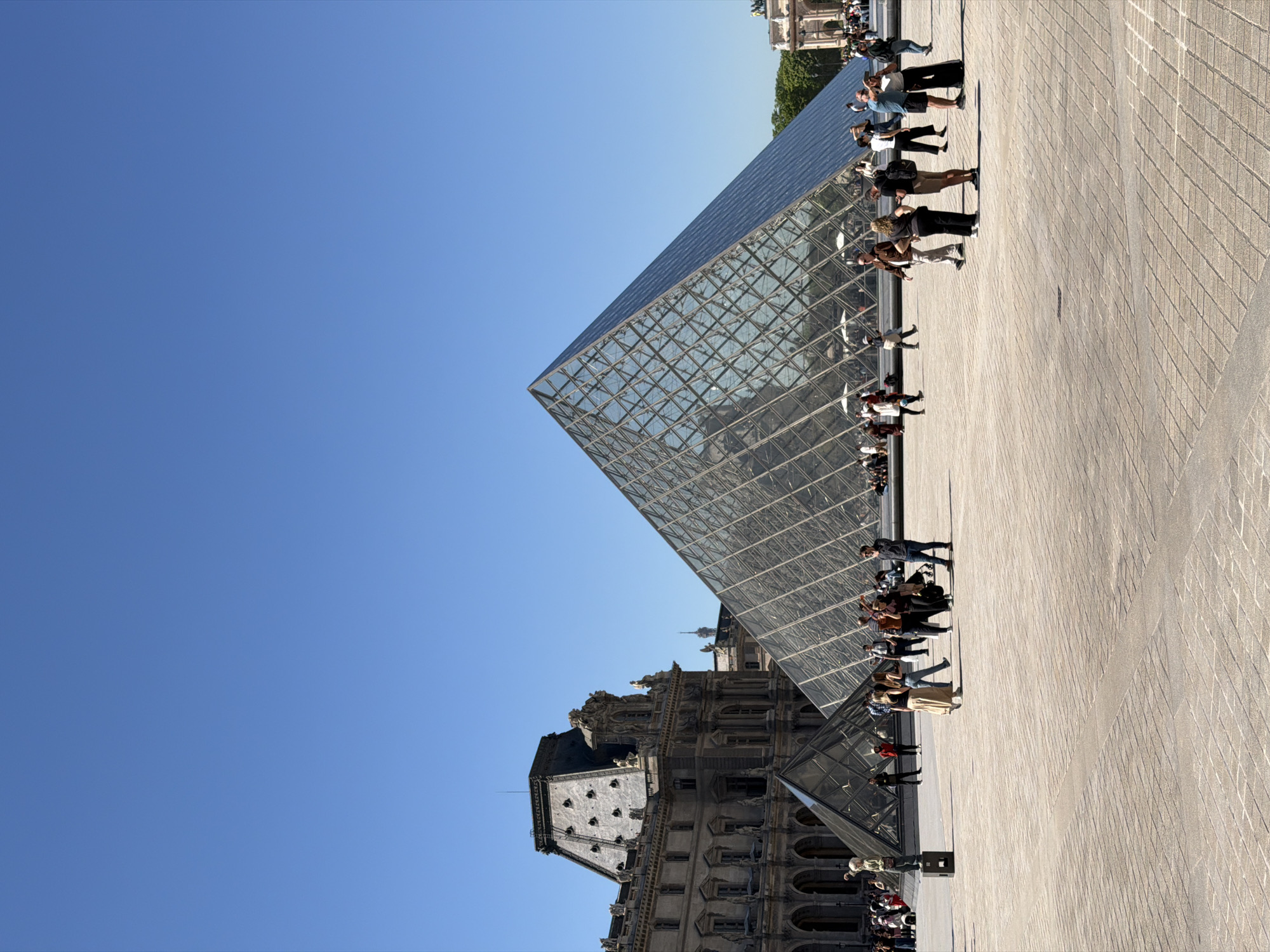 The image captures the Louvre Pyramid in Paris, France, with people positioned around it. The pyramid stands against a clear blue sky. Adjacent to the pyramid is the Louvre Palace, showcasing its architectural features. The foreground is a paved area. People are seen walking and standing, some interacting with the environment, while others are presumably taking photographs. The overall scene is typical of a tourist attraction.