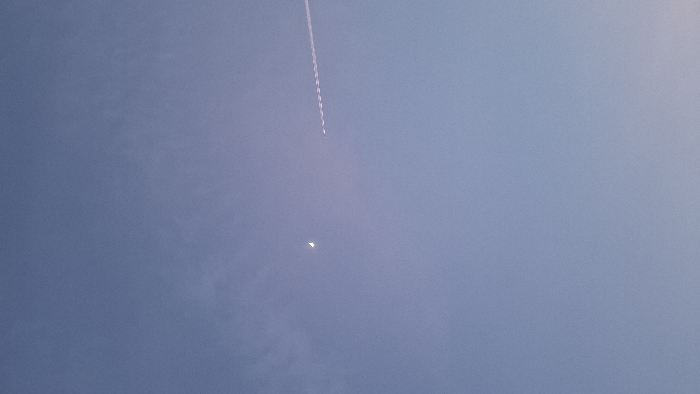 The scene, observed from Boreti, Montenegro, depicts an expansive view of the twilight sky. The upper portion of the sky exhibits a pale blue hue, transitioning into a lighter, subtly purple gradient towards the lower right. A distinct, slender crescent moon is positioned in the upper-left quadrant. To the right, a long, linear aircraft contrail extends diagonally across the upper-right section of the frame, originating from the right edge and moving towards the mid-left. The contrail is white and appears to be segmented or broken into distinct dashes. No other celestial bodies, aircraft, landmasses, structures, individuals, or associated actions or interactions are visible.