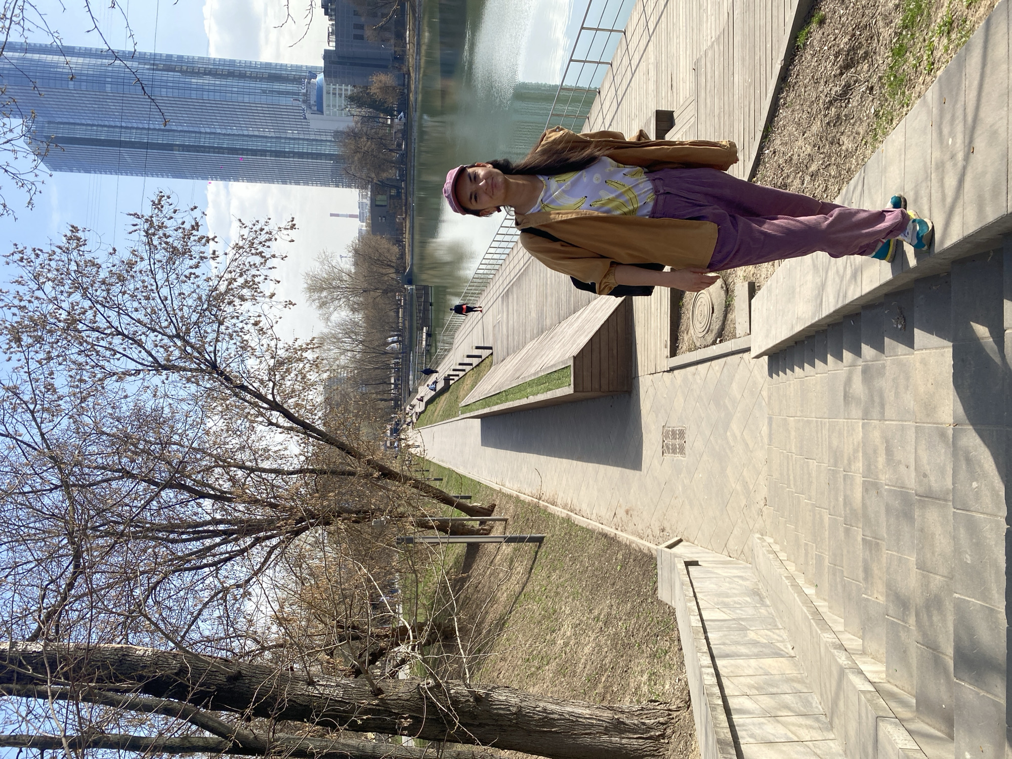 A young woman is standing on a sidewalk in Khimki, Russia. She is wearing a tan jacket, a purple baseball cap, a white T-shirt with yellow bananas, and purple corduroy pants with bright-colored sneakers. In the background is a river and a cityscape including a tall glass skyscraper. There is a wooden boardwalk next to the river with benches. Trees surround the walkway and the grass is dry.