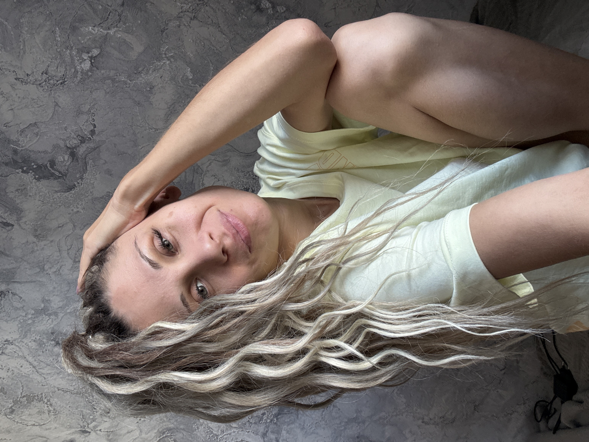 A woman with blonde and brown ombre hair tied in a high ponytail is posing for a selfie. Her hand is resting on her head, and she is looking towards the camera with a slight smile. She is wearing a light yellow t-shirt. The background appears to be a textured grey wall, suggesting an indoor setting. There is no visible indication that this scene is located in Rusanovo, Russia.