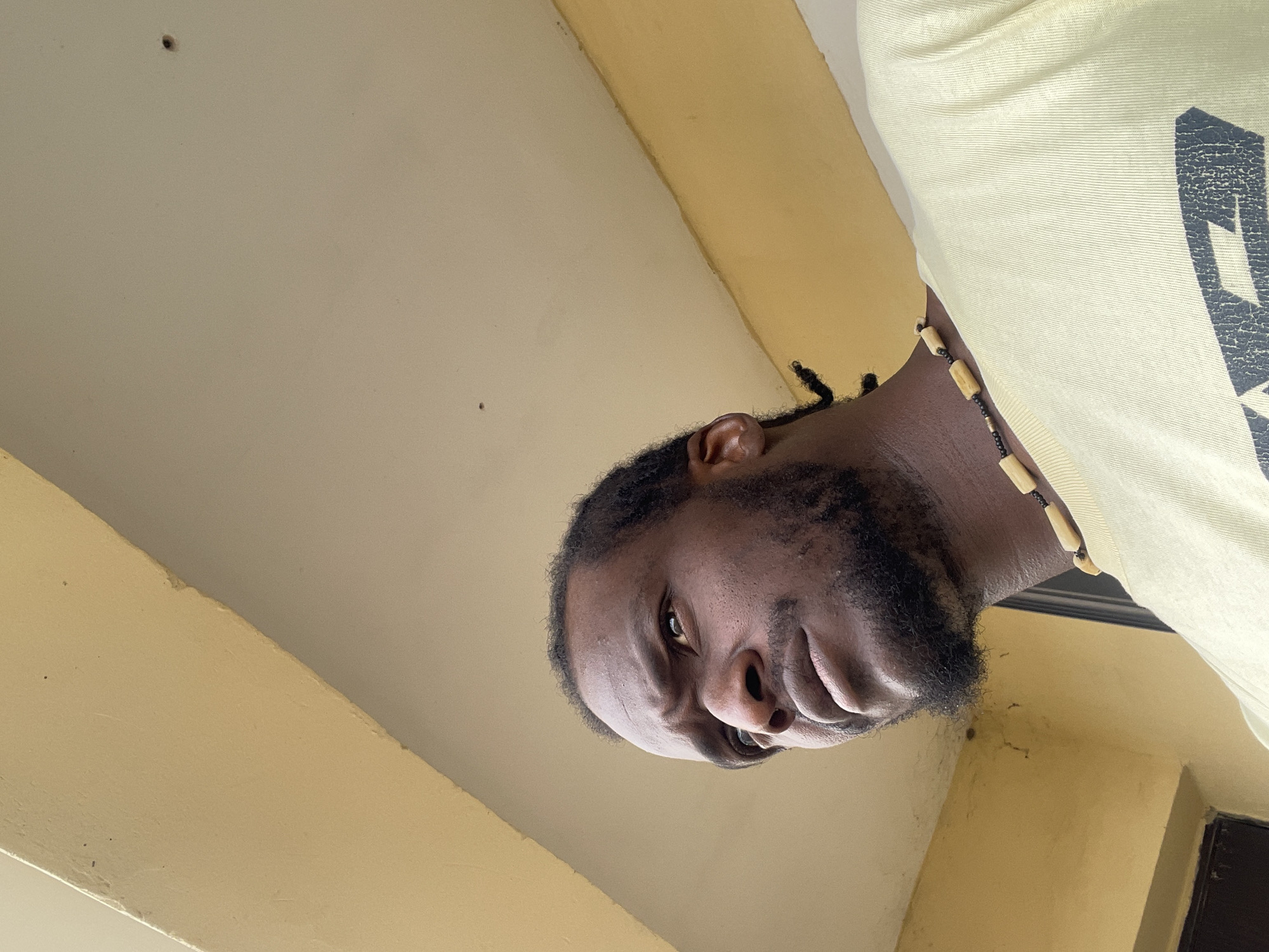 The image captures a medium shot of a Black man with dark skin, a beard, and dark hair styled in braids. He is wearing a light yellow t-shirt and a necklace made of wooden beads. The man is looking upwards and to his left with a thoughtful or concerned expression. The background is a muted yellow wall and a white ceiling, suggesting an outdoor or semi-outdoor location. The lighting indicates it is daytime. The environment appears to be a simple, possibly residential or urban, setting. The texture of the wall shows some signs of age or weathering. The provided hints show close-ups of the man's face and clothing, as well as details of the yellow wall and white ceiling. It is not possible to determine the exact location within Abuja, Nigeria, from the image alone.