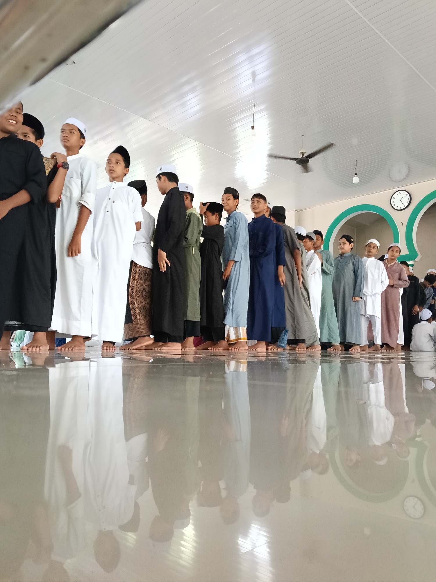 A ground-level, medium shot captures a line of young boys, mostly in light-colored traditional clothing and caps, barefoot, standing in a row on a shiny, reflective floor. Their reflections are visible on the floor. The boys are of varying heights and skin tones, and their clothing consists of different styles and colors of robes and tunics.
Behind the boys, a wall with a green and white arched design and a clock are visible. A ceiling with fluorescent lighting and a ceiling fan is above them. The overall lighting is bright and seems to be from the ceiling lights. The scene appears to be inside a mosque in Lhokseumawe, Indonesia.