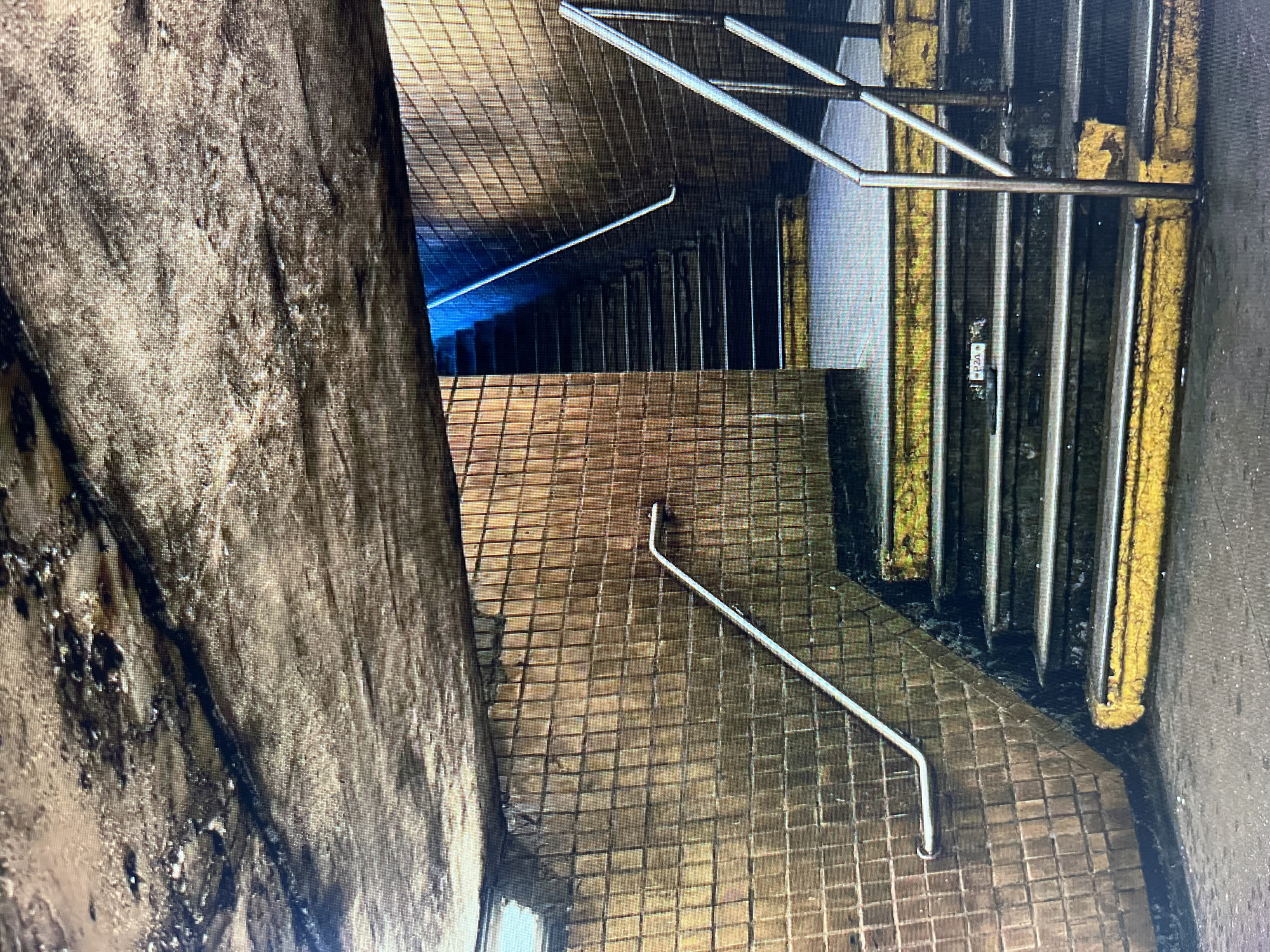 An interior shot of a subway staircase shows weathered tiled walls and a rough concrete ceiling. The staircase itself is made of concrete steps with yellow and black painted risers and edges. Metal handrails line both sides of the stairs. A sign that says "F20" is visible on one of the steps. The overall scene suggests a grimy and utilitarian urban environment.