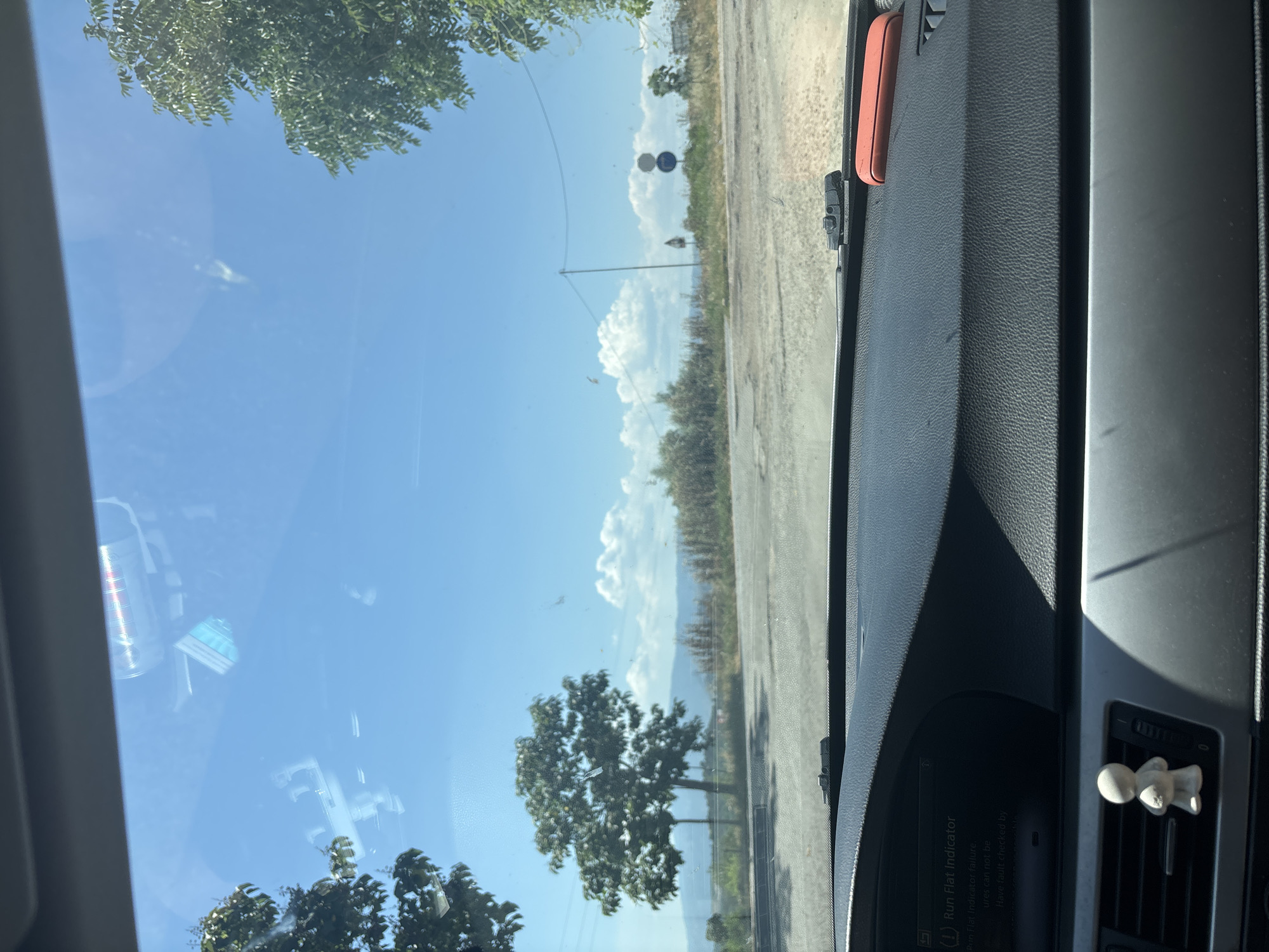 The image captures a static scene from within a vehicle in Marikostinovo, Bulgaria, looking outward through the windshield. The vehicle's dark, textured dashboard occupies the lower portion of the frame. A digital display on the dashboard indicates "(!) Run Flat Indicator" with text detailing a "Run Flat Indicator failure." A small, white, stylized humanoid figurine is affixed to an air vent on the left side of the dashboard, while an orange rectangular object rests on the far right. Reflections of interior elements, including what appears to be a metallic can and other indistinct items, are visible on the windshield glass.
Outside, under a clear blue sky dotted with scattered white cumulus clouds, an unpaved or gravely road surface is visible in the immediate foreground, leading into the mid-ground. Green foliage from trees partially frames the view on the left and right. A prominent, bushy green tree stands centrally to the left of the road. Further right, a utility pole with overhead wires is present, along with more scattered dry vegetation. Two circular regulatory road signs, one predominantly blue with a red diagonal stripe and the other blue and white, are visible in the mid-distance on the right side of the road. In the far background, faint outlines of rolling hills or mountains are discernible beneath the horizon line of more distant vegetation. No people are visible.