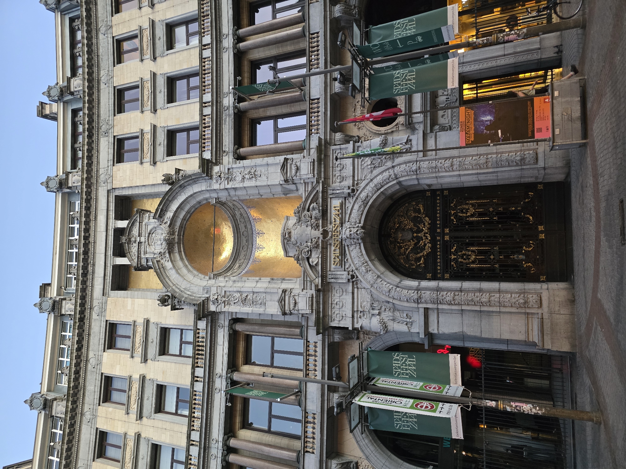 The image shows the exterior of the historic Stadsschouwburg building in Antwerp, Belgium, a grand architectural structure with ornate stone carvings and classical design elements. The building features a large, intricately decorated arched entrance with golden detailing. Above the entrance, there is a semicircular niche filled with golden mosaic tiles. Several tall, green banners with white lettering hang from poles in front of the building. One banner reads "STADS FEEST ZAAL," and another appears to mention "ORIENTAL." A red and white flag is also visible on a pole. In front of the main entrance, there is a metal fence and a sign advertising an event titled "ZOMER MALAM NIGHT." The paving stones in front of the building are arranged in a patterned walkway. The sky above is a clear, pale blue, indicating it is either early morning or late evening.