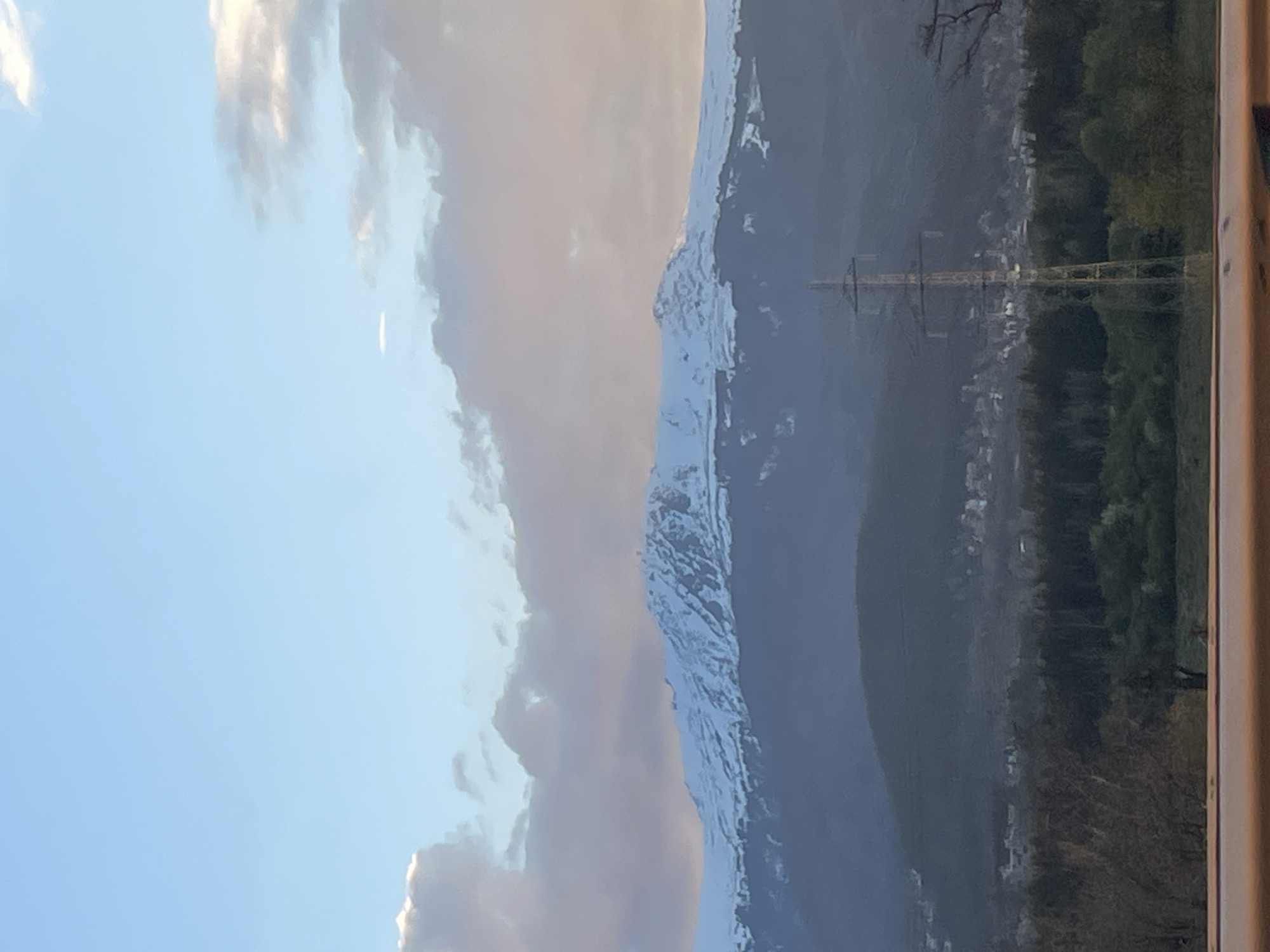 The image presents a wide, vertically oriented landscape view, likely captured from a window, as indicated by a partial frame visible on the far right.
The scene is dominated by a majestic mountain range stretching horizontally across the middle of the frame (when rotated correctly). These mountains feature prominent snow-capped peaks, suggesting a high altitude or cold climate. Above the mountains, a vast expanse of sky is visible, transitioning from clear blue on the left to an area with scattered, soft white and grey clouds on the right. One of the cropped sections highlights a thin, bright white line in the blue sky, possibly a distant airplane contrail.
Below the mountain range, in the mid-ground and foreground, lies a valley or lower terrain. This area is characterized by dense tree cover, appearing as a forest with varying shades of green and brown foliage. Nestled within this landscape, a small settlement or village is discernible, with several buildings scattered among the trees and foothills. A prominent metallic electricity pylon stands tall in the foreground, its intricate structure silhouetted against the mountain and valley background.
The lighting suggests either early morning or late afternoon, with soft illumination on the clouds and snow-capped peaks, while parts of the valley and mountainsides are in shadow or appear slightly hazy. No people or specific activities are depicted. No text is visible in the image.