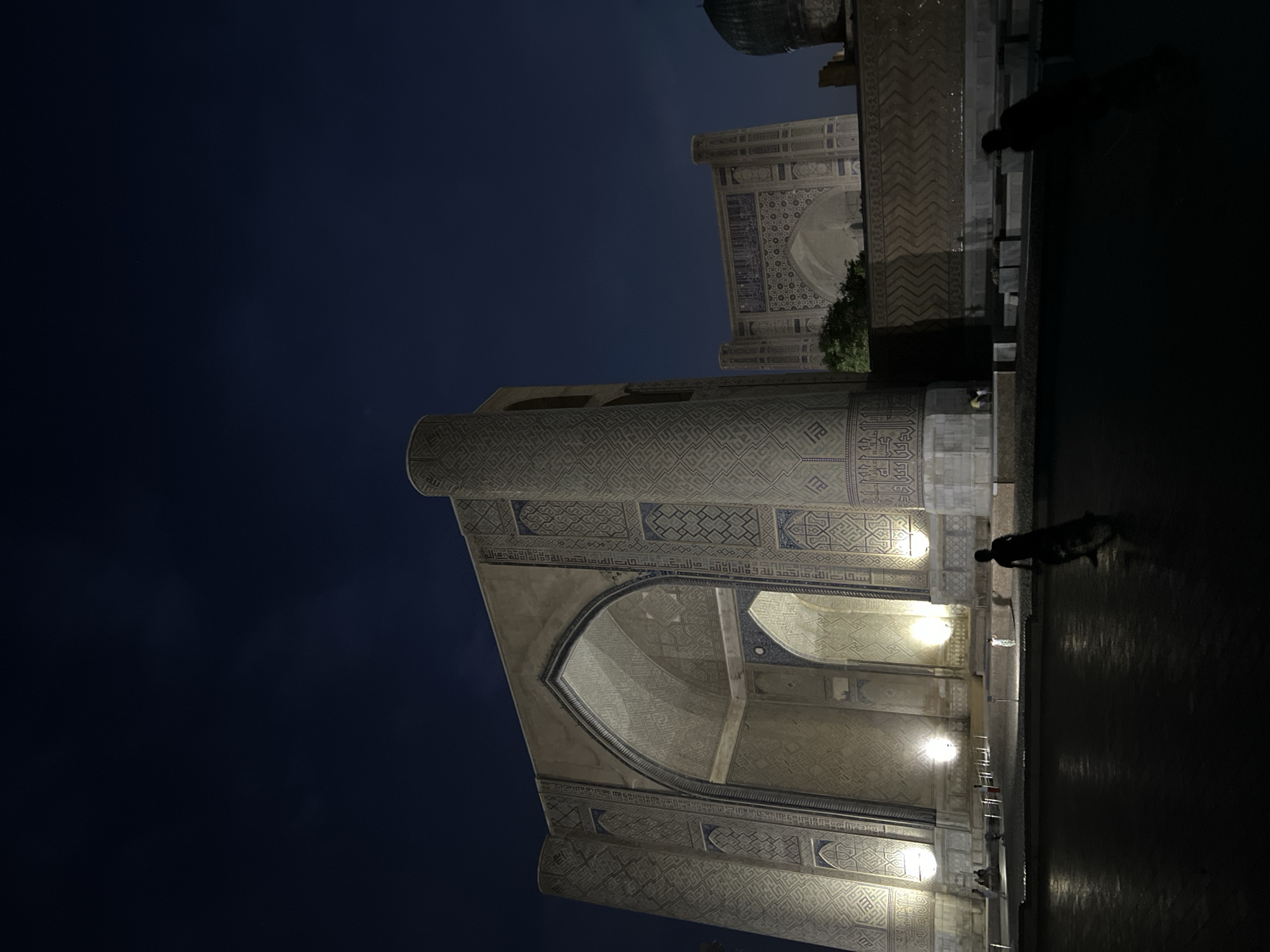 The image depicts an exterior view of an ancient architectural complex at night. The scene is dominated by a grand, intricately decorated building, likely a mosque or madrasah, illuminated by artificial lights. The architecture features elaborate geometric patterns and Arabic calligraphy etched into the facade, showcasing intricate tilework in shades of blue and beige against a light stone background. Large arched doorways and towering minarets suggest a significant historical and religious site.
In the foreground, a dark, reflective surface, possibly a pool of water or wet paving, mirrors the illuminated structures and the deep indigo night sky. The sky is clear, with no visible clouds, and the overall atmosphere is serene and majestic, amplified by the nighttime setting.
Several silhouetted figures are present, suggesting that the location is accessible to visitors even after dark. One figure is seen standing near the water's edge, appearing to be observing the architecture. Another figure is closer to the building, possibly walking or pausing. The presence of people adds a sense of scale and life to the monumental structures.
The time of day is clearly night, indicated by the dark sky and artificial lighting. The weather appears calm, as suggested by the clear sky and the undisturbed reflections on the ground. The architectural style and the use of calligraphy point towards a location with rich Islamic heritage, consistent with the provided context of Samarkand, Uzbekistan. The overall impression is one of awe and historical reverence, a timeless beauty preserved under the cover of darkness. No legible text is discernible in the image.