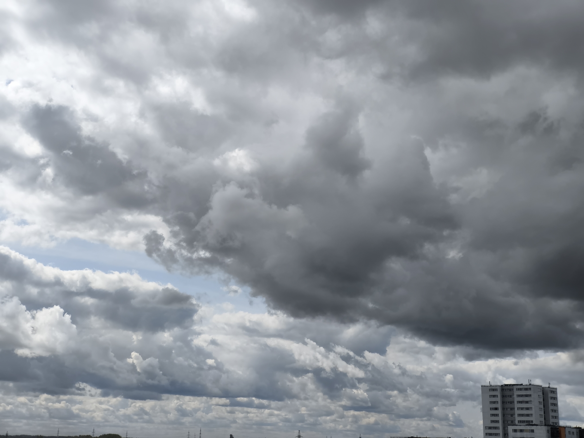 The image captures a view of a cloudy sky with a large, modern apartment building on the right side of the frame. The sky is dominated by heavy, dark grey clouds, suggesting impending rain or a storm. Some lighter clouds are visible in the upper left portion of the image, with a small patch of blue sky peeking through. The apartment building is tall and white with many windows, characteristic of urban architecture. The lower part of the image shows a distant horizon line with some faint structures that could be other buildings or industrial elements, and what appear to be power lines or transmission towers. The overall impression is one of a dramatic sky over an urban landscape in Tallinn, Estonia, with the focus on the atmospheric conditions and the imposing presence of the building.