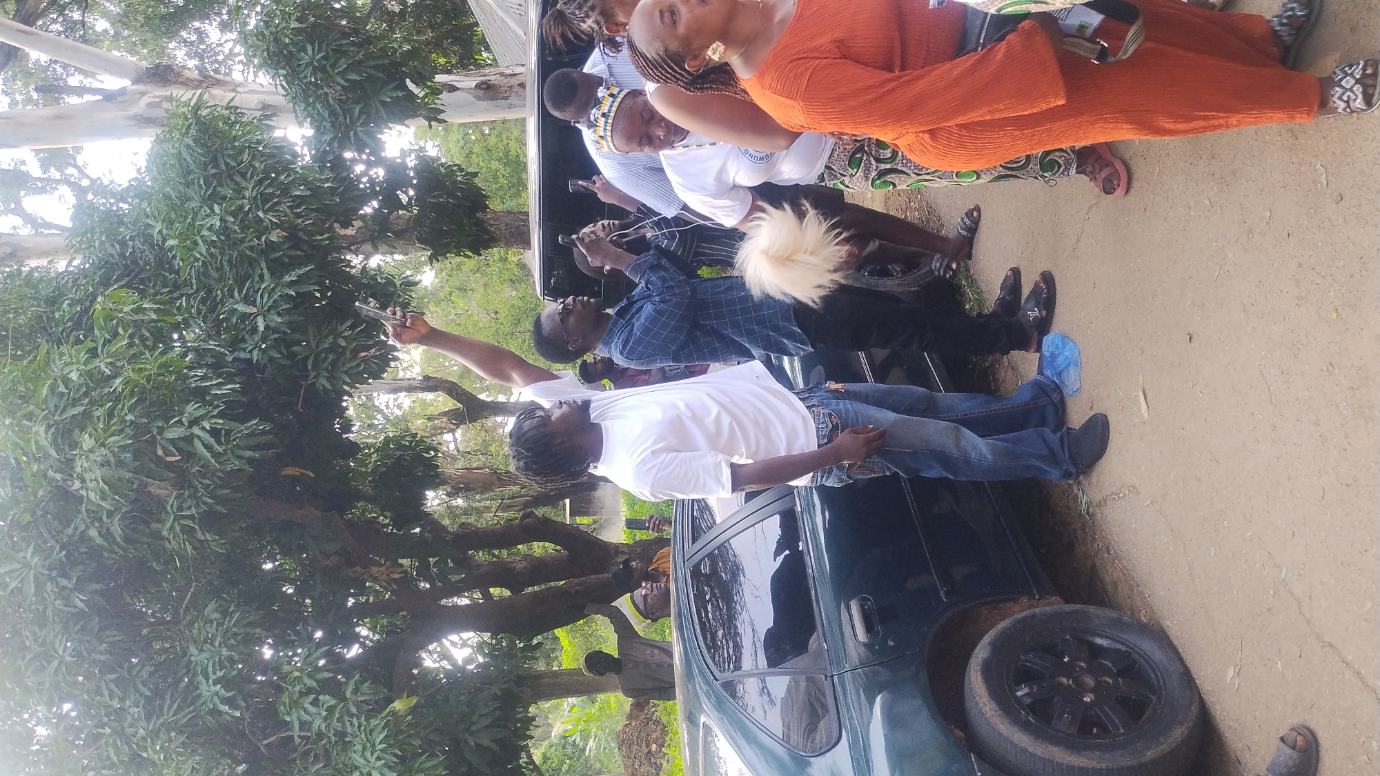 The image depicts an outdoor scene, likely in Jos, Nigeria, with a group of people gathered near a dark-colored sedan. The ground is a dirt road, and trees with lush green foliage dominate the background. Sunlight filters through the leaves, casting dappled shadows.
In the foreground, several individuals are standing. A man on the left, wearing a white t-shirt and blue jeans, is holding a smartphone up, possibly taking a photo or video. To his right, another man in a blue checkered shirt is also holding a smartphone. To the right of these men, a woman in an orange outfit is partially visible, and further to the right, another woman in a white t-shirt and patterned skirt is visible. The woman in the orange outfit appears to be speaking.
Behind the group, a dark SUV is parked. Further in the background, other individuals are visible, some of whom appear to be wearing traditional attire. The overall atmosphere suggests a gathering or event, with people documenting the occasion. The presence of the car, the people, and the trees are consistent with a setting in a Nigerian city like Jos.