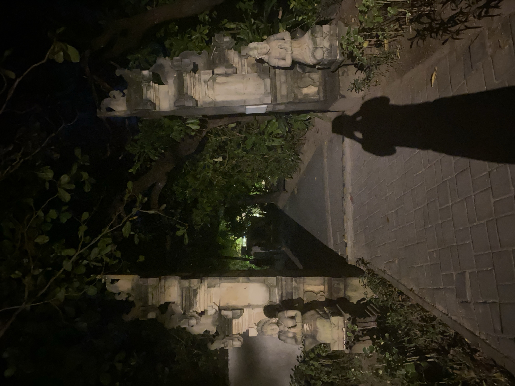 A nighttime image in Kuta Selatan, Indonesia depicts a stone pathway leading between rows of ornate, carved stone structures, potentially gates or decorative walls. The structures are weathered and surrounded by dense vegetation. A long shadow, likely cast by a person holding the camera, stretches across the brick pathway. The light is dim, with a few distant lights visible between the structures.