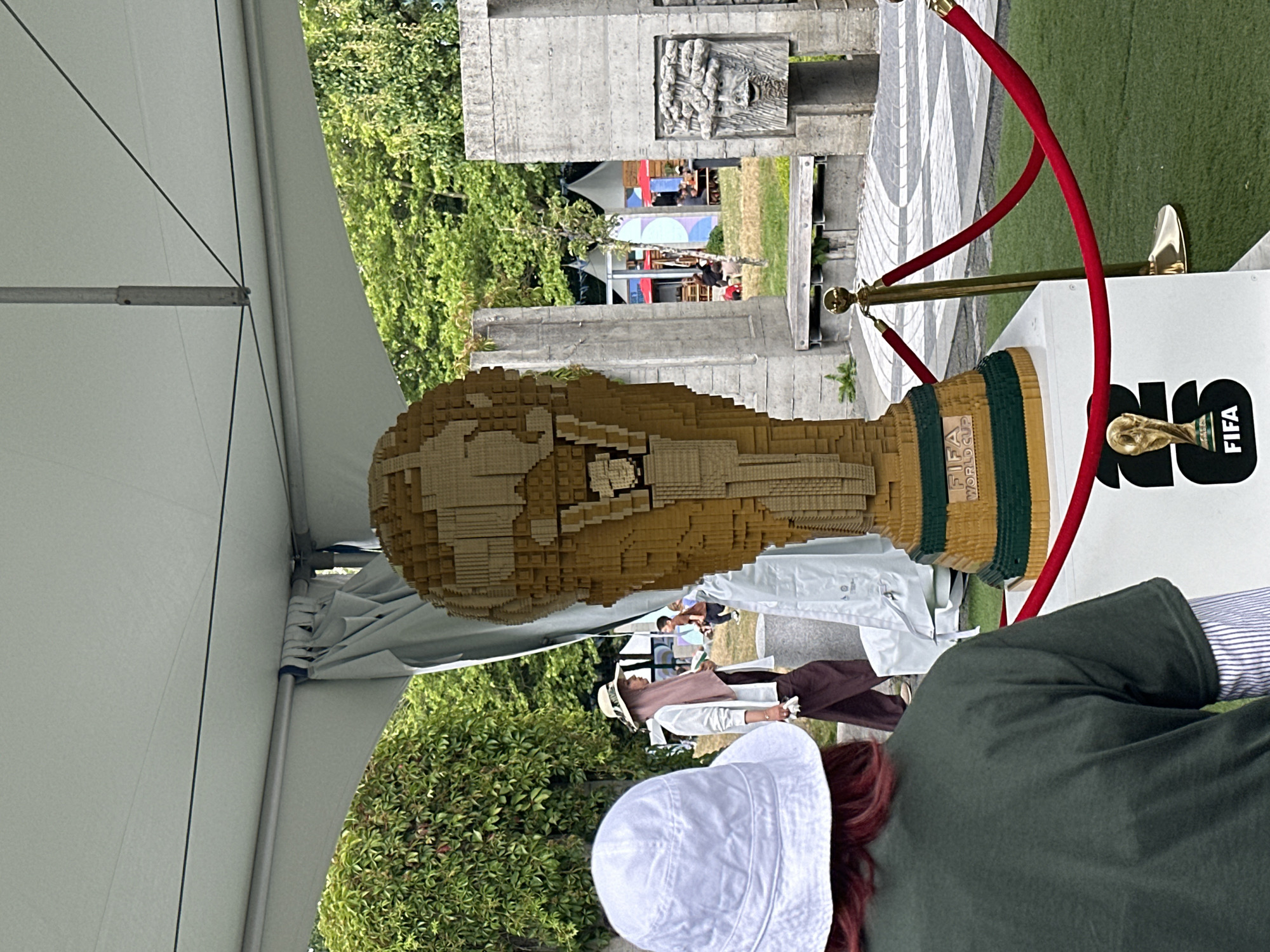 In a park-like setting in Vancouver, Canada, a large, golden FIFA World Cup trophy, constructed from LEGO bricks, is displayed under a white tent. The trophy rests on a white pedestal that features the FIFA World Cup logo. A red rope barrier, held by gold stanchions, surrounds the display, preventing closer access.
In the foreground, the back of a person wearing a dark green t-shirt and a white bucket hat is visible, facing the trophy. To the left, another person wearing a light-colored hat and a pink hijab walks away from the camera, dressed in a white shirt and dark pants. In the background, several concrete structures with carved faces and architectural elements are visible, along with lush green trees and foliage. Further back, tents and people can be seen, suggesting a public event or exhibition. The ground is covered with green turf and a paved area with a patterned design.
