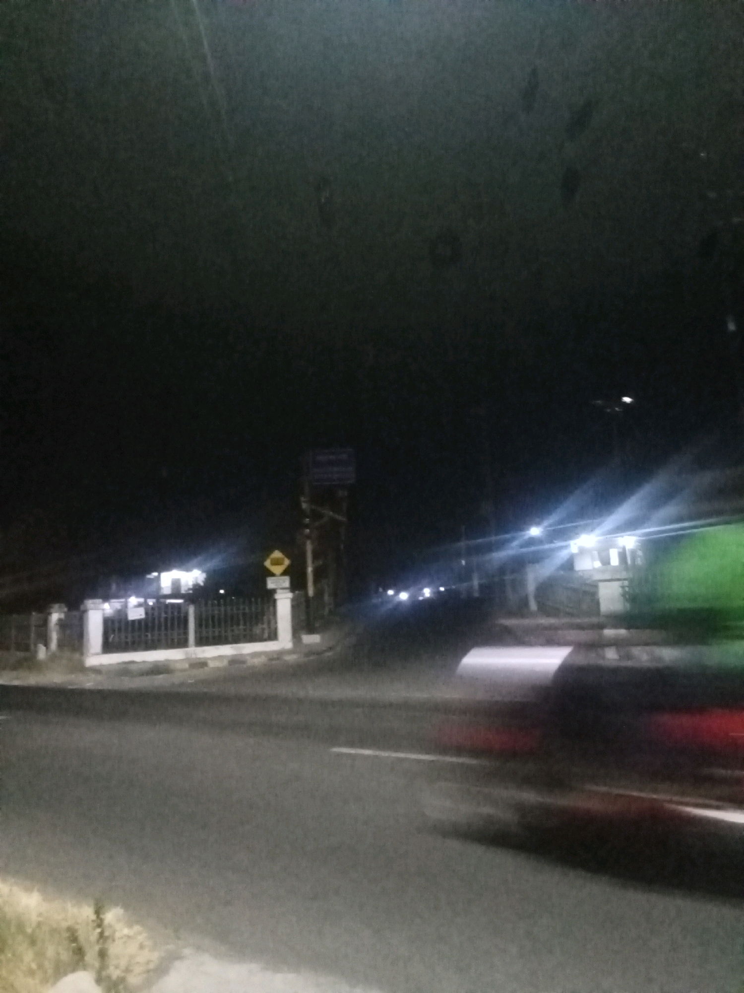 The image depicts a nighttime scene on a road in Sumber Mulyo, Indonesia. The foreground shows a paved road with white lane markings. On the left side of the road, there is a strip of vegetation. In the mid-ground, there is a white fence with a metal railing, behind which is a building with bright lights illuminating the area. A yellow diamond-shaped sign with a black symbol is visible on a pole to the right of the fence. Further down the road, there are more buildings and streetlights. A vehicle is blurred in motion on the right side of the road, suggesting it is traveling at a significant speed. The overall lighting is dim, with artificial lights from buildings and streetlights creating strong highlights and shadows. The sky is dark.