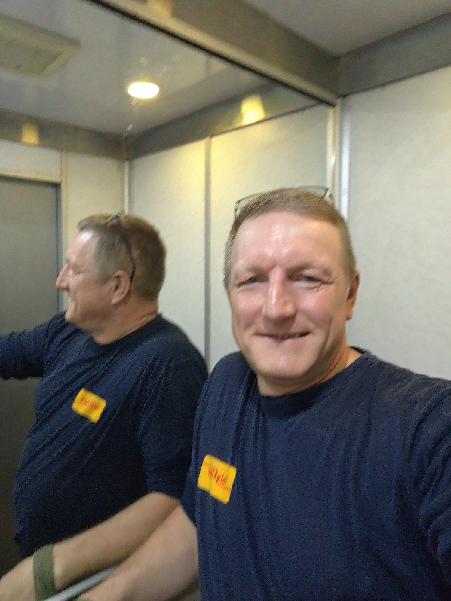 The image depicts a man taking a selfie in what appears to be an elevator. The man, in his late 40s to early 50s, has fair skin, short brown hair, and is wearing glasses pushed up on his head. He is smiling at the camera. He is wearing a dark blue t-shirt with a yellow rectangular patch on the left breast that says "Pakadi". In the reflection of the elevator's mirrored wall, another man in a similar t-shirt is visible, turned away from the camera and looking to his left. The elevator interior has light-colored walls and a light-colored ceiling with a circular fluorescent light fixture. There is a metal handrail visible in the reflection. The overall environment suggests an interior space within a building, possibly a workplace, in Petah Tikva, Israel, given the presence of the "Pakadi" patch, which is a known Israeli company.