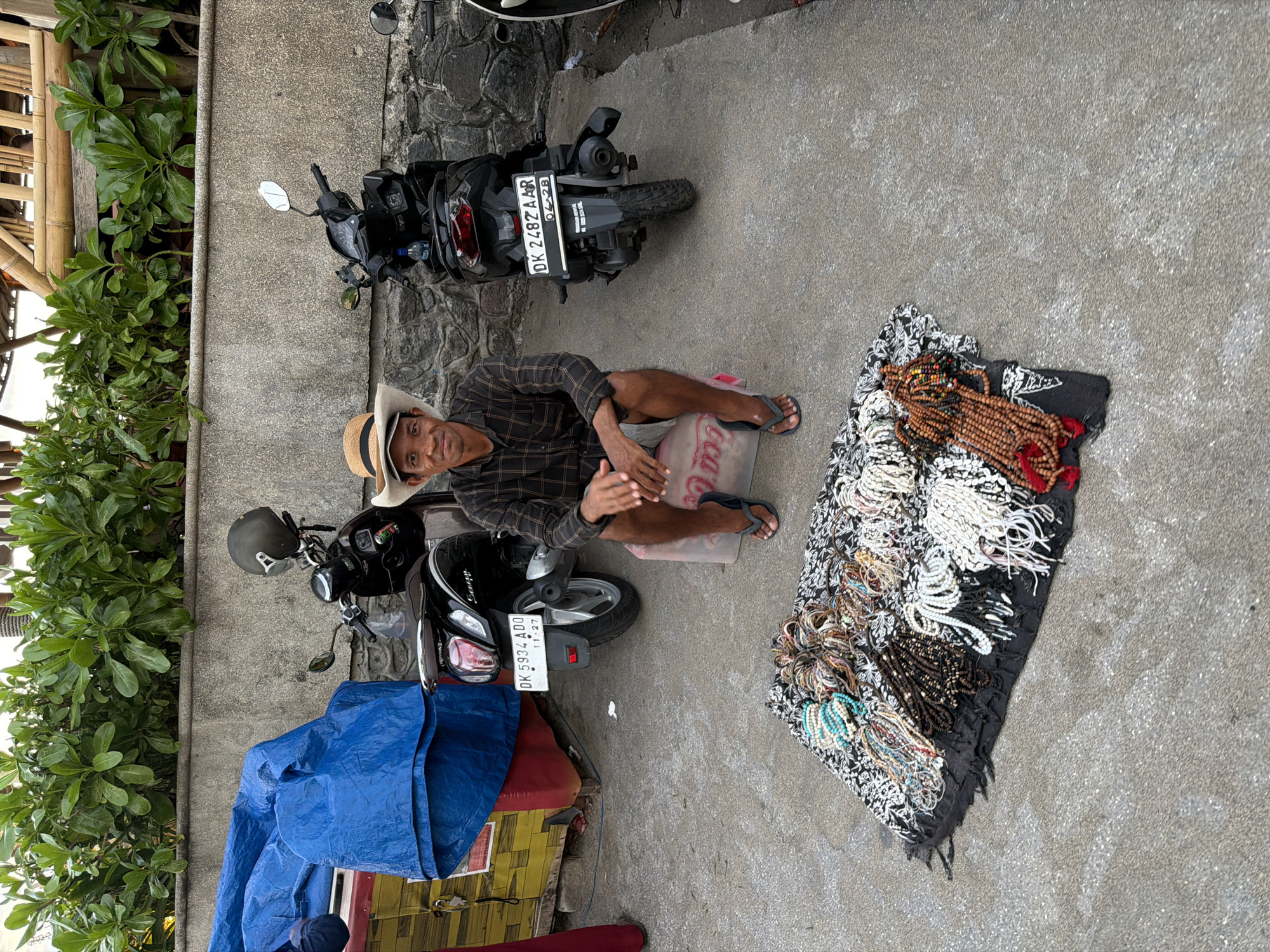 The image depicts a street vendor sitting on the ground in North Kuta, Indonesia, with his merchandise displayed on a patterned cloth. He is a man wearing a checkered shirt, shorts, and a straw hat. His legs are crossed, and he appears to be interacting with someone, possibly showing them his wares or waiting for customers.
The vendor's merchandise consists of numerous beaded necklaces and bracelets, displayed on a black and white patterned cloth. The beads are made of various materials, including wood, stone, and what appears to be shell or plastic, in a range of colors and sizes.
Behind the vendor, two scooters are parked. One is a black scooter with a license plate that reads "DK 2482 Z AAR". The other scooter, partially covered by a blue tarp, has a license plate that reads "DK 5934 ADD". To the left of the scooters, there is lush green foliage growing next to a textured stone wall and some bamboo structures.
The scene appears to be outdoors, possibly on a sidewalk or a small roadside stall. The ground is concrete. The lighting suggests it is daytime, and the weather seems to be overcast or partly cloudy, as there are no harsh shadows. The vendor's expression is neutral, and he seems relaxed. There is no visible text on the merchandise, but the license plates on the scooters are legible.