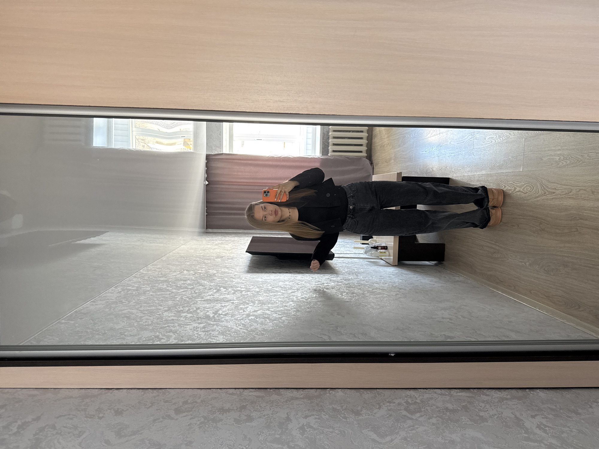 A young woman is captured in a mirror selfie, standing indoors in what appears to be a modern room, possibly in Chaikovsky, Russia. She has long, straight blonde hair and is dressed in a black jacket, dark jeans, and light-colored slip-on shoes. She holds an orange smartphone in her right hand, using it to take the photograph of her reflection.
The room, visible in the mirror's reflection, is brightly lit by natural light streaming through a large window, which is partially covered by a light-colored blind or curtain. Below the window, a white radiator is visible. The floor inside the reflected room is light wood laminate, showing a distinct grain pattern. To her left, a dark, low-profile piece of furniture, possibly a stand or bench, is present, along with a smaller, lighter-colored shelf holding various small objects. The mirror itself has a visible wooden frame at its top and bottom edges (in the original image orientation). The floor directly in front of the mirror, outside the reflected space, is a patterned grey texture. The time of day appears to be daytime due to the natural illumination. The woman displays a calm, neutral expression. No visible text is discernible within the image.