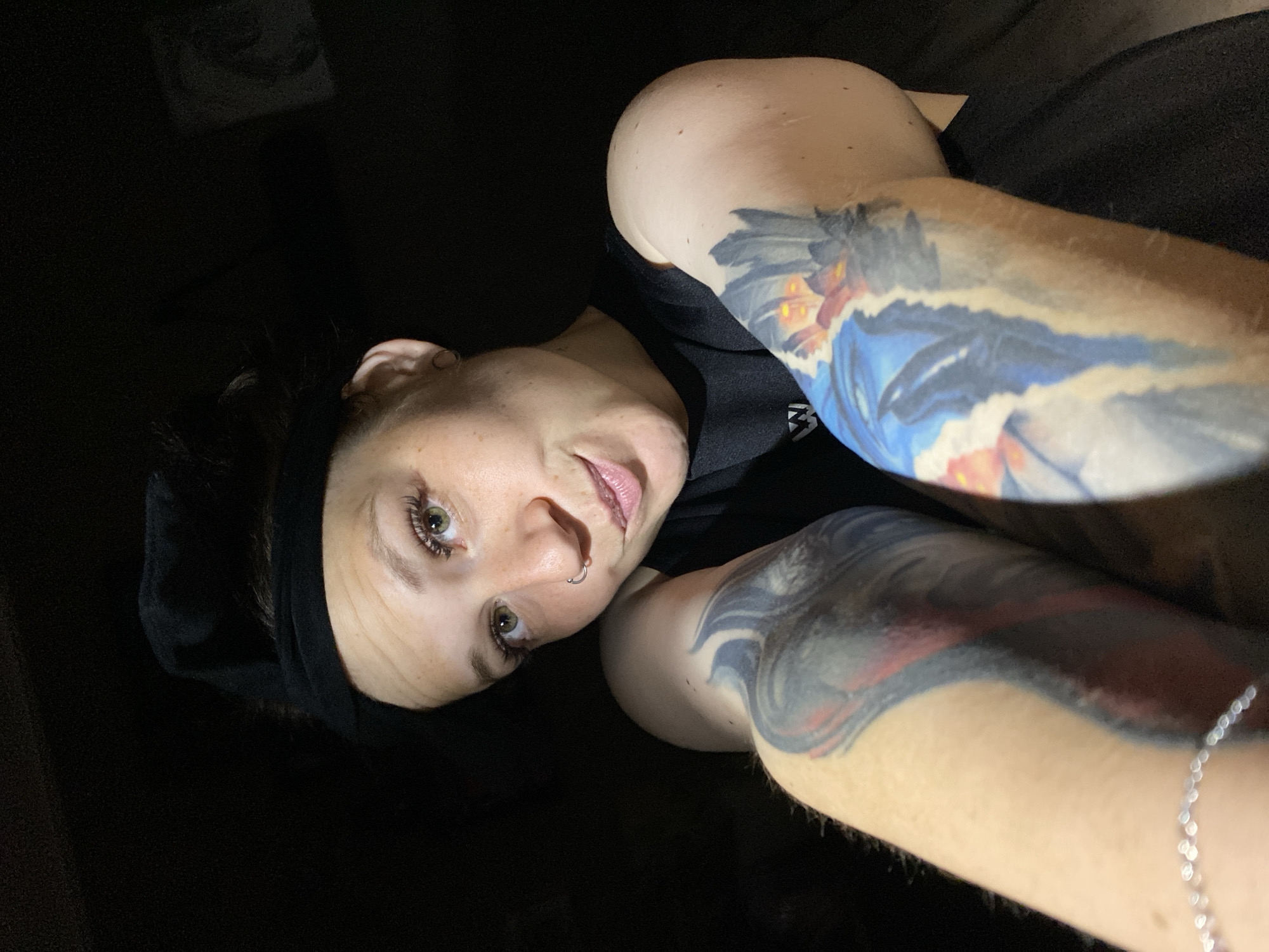 In a dimly lit indoor setting, a young Caucasian woman with light brown hair pulled back and a black headband is shown in a close-up selfie. She has green eyes, a nose ring, and is wearing a black t-shirt. Her arms are adorned with colorful tattoos, one on her left arm featuring shades of grey and red, and the other on her right arm with blues, oranges, and blacks. A thin silver bracelet is visible on her left wrist. The background is dark, with a faint outline of a framed image on the upper right. The image does not provide any specific information that would allow for the localization of the scene within the city of Chaikovsky, Russia.