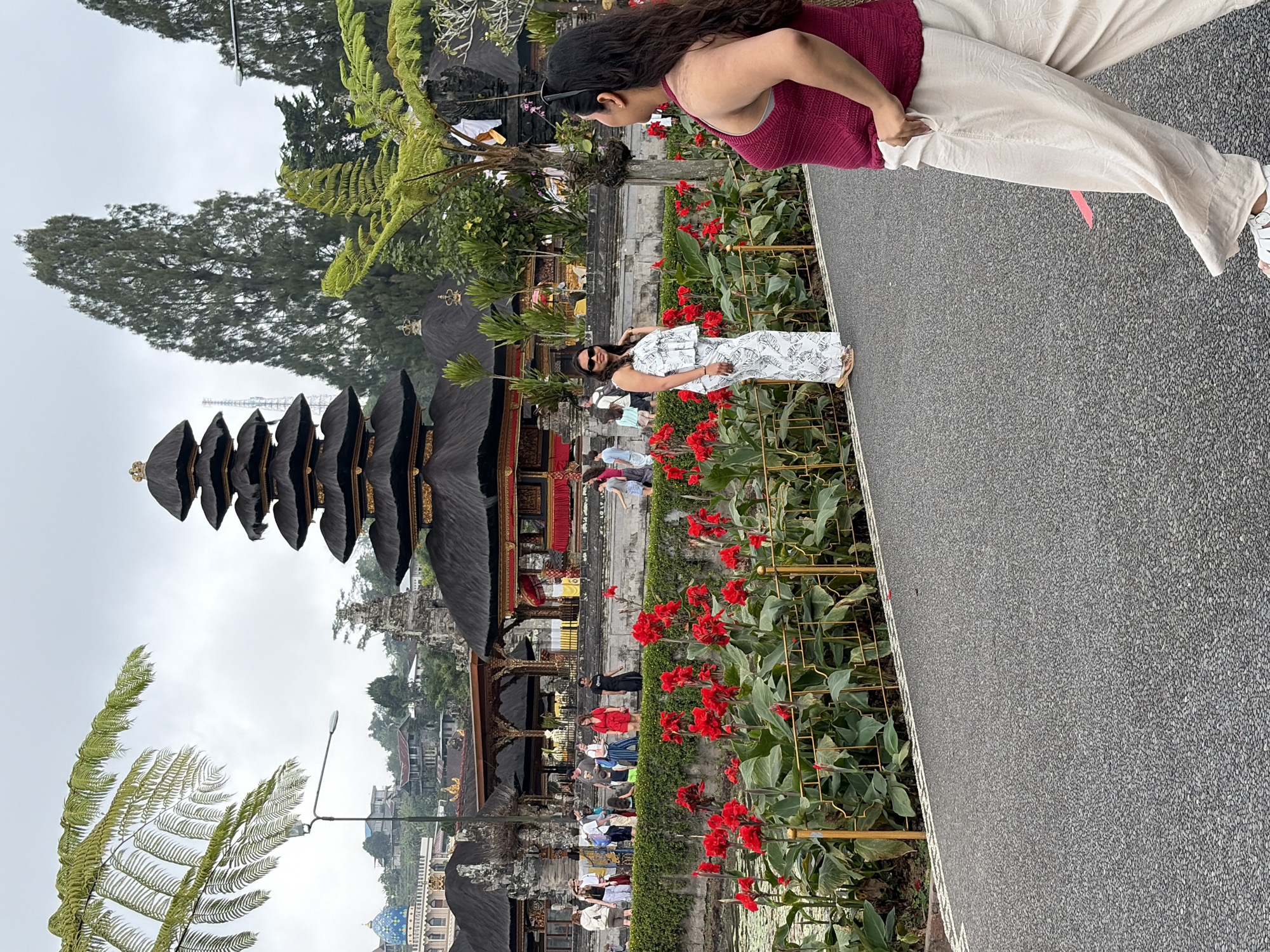 The image depicts the Pura Ulun Danu Bratan temple complex in Baturiti, Indonesia, under an overcast sky. In the foreground, a woman wearing a maroon tank top and loose cream-colored pants walks away from the camera, her right hand in her pocket. To her left, on a paved path, a woman in a white and green patterned outfit stands and looks towards the camera, her left hand on her hip. Behind them, a vibrant bed of red flowers lines a low green hedge, separated from a body of water by a golden fence. In the midground, several other tourists are visible, walking and observing the temple architecture. The iconic multi-tiered pagoda of the temple stands prominently, its dark roof contrasting with the surrounding greenery and a distant blue-domed building. Tall trees and foliage frame the scene, with a fern frond arching into the top left corner of the frame.