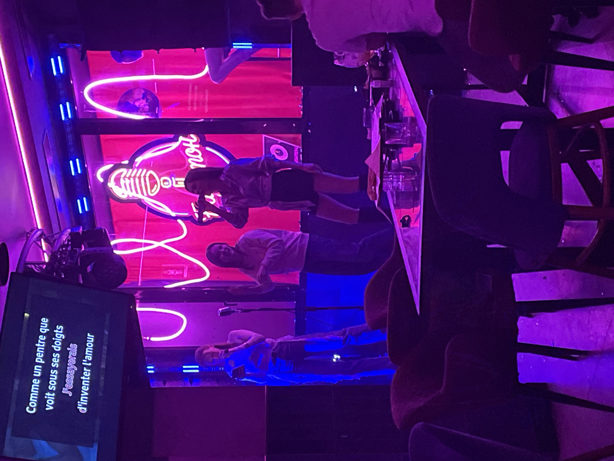The image shows a karaoke bar, likely in Perm, Russia, given the Cyrillic text on the screen. The atmosphere is dimly lit with vibrant pink and blue neon lights. Patrons are engaging in karaoke, with lyrics displayed on a screen. People are visible both singing and watching, suggesting a lively social atmosphere. Tables are set with drinks, and chairs are arranged around them, indicating a place for gathering and enjoying the entertainment. The decor features neon signs and light displays, typical of a modern karaoke establishment.
