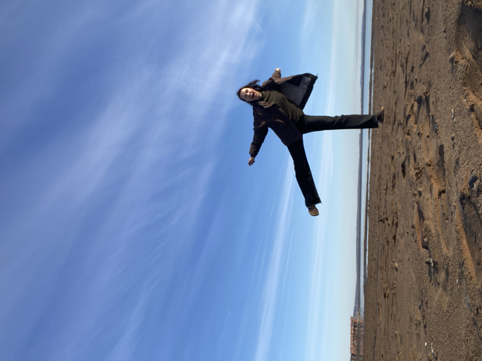A full-body, low-angle photograph captures a woman in mid-air, seemingly jumping or floating, against a vast, bright blue sky. She is dressed in a dark jacket, dark top, and dark pants, with light brown shoes. Her arms are slightly spread, and her legs are apart, conveying a dynamic sense of playful motion. Her face displays an open-mouthed smile, indicating joy or excitement.
The setting appears to be a wide-open outdoor space, likely a shoreline or beach next to a body of water, consistent with the Kama River in Chaikovsky, Russia. The ground, visible across the bottom and right side of the image, is dark brown, textured, and appears to be sand or mud, with small pebbles and tire tracks. A calm body of water stretches to the horizon, reflecting the bright sky. In the far distance along the water, faint structures or buildings are discernible. The sky is clear, a brilliant blue, with soft, wispy white clouds scattered across it, suggesting a sunny daytime. There is no visible text in the image.