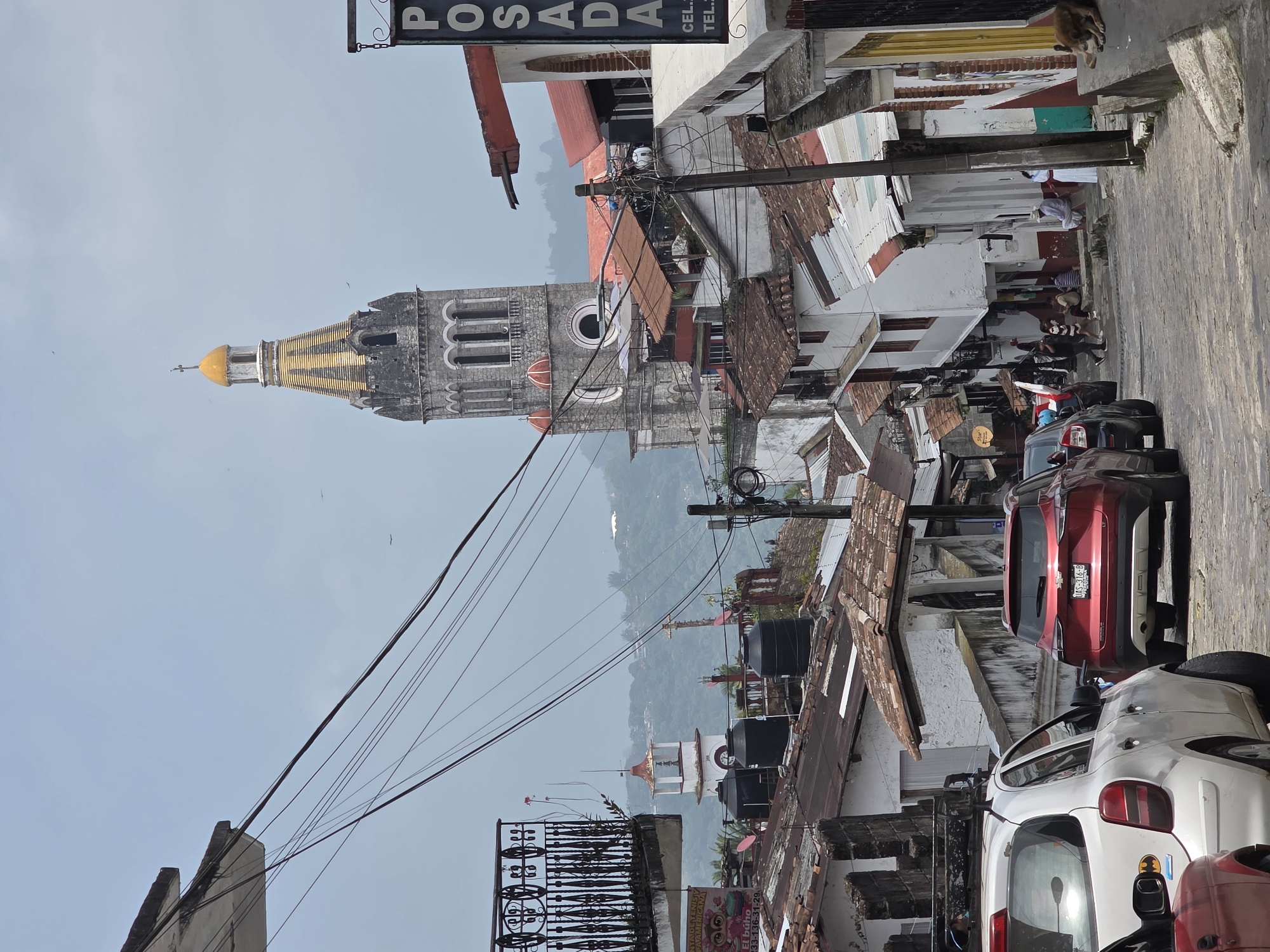 The image depicts a street scene in Cuetzalan, Mexico, looking down a narrow street lined with buildings. The most prominent feature is the tower of the Iglesia de San Francisco de Asís, a notable landmark of the town. The tower is stone, with a gold-colored dome.
Several cars are parked on the right side of the street, with a red SUV and a white compact car visible. People are present on the sidewalk, some sitting and others standing. Overhead, a dense network of electrical wires stretches across the sky. Signs for "POSADA" and "El búho" are visible, along with a license plate number "UAS-316-B" on one of the cars. The buildings are a mix of white and red, with tiled roofs. In the background, green hills can be seen. The overall impression is of a typical street scene in a historic Mexican town.