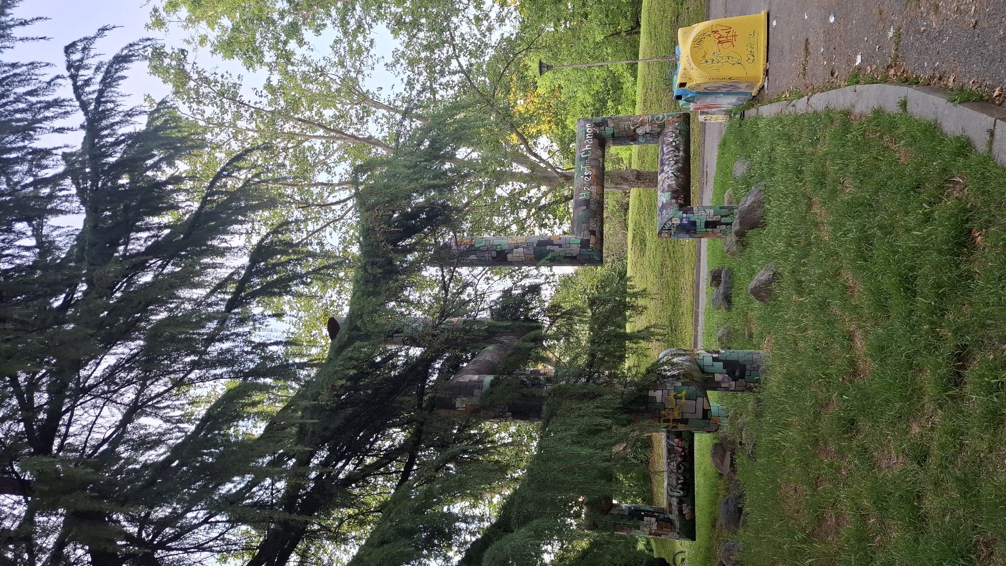 In the city of Sofia, Bulgaria, an outdoor art installation is situated in a park, likely a public space given the presence of a trash receptacle. The sculpture, composed of angular, interlocking geometric shapes, is covered in a mosaic of colorful tiles and graffiti. It is surrounded by green grass, scattered rocks, and the branches of a large coniferous tree in the foreground. In the background, lush trees and a bright sky suggest a pleasant day. A yellow trash can decorated with yellow graffiti and a white drawing of a face is visible to the right of the sculpture, next to a paved path. The sculpture itself appears to have text, possibly "Here's to Change," inscribed on one of its segments.