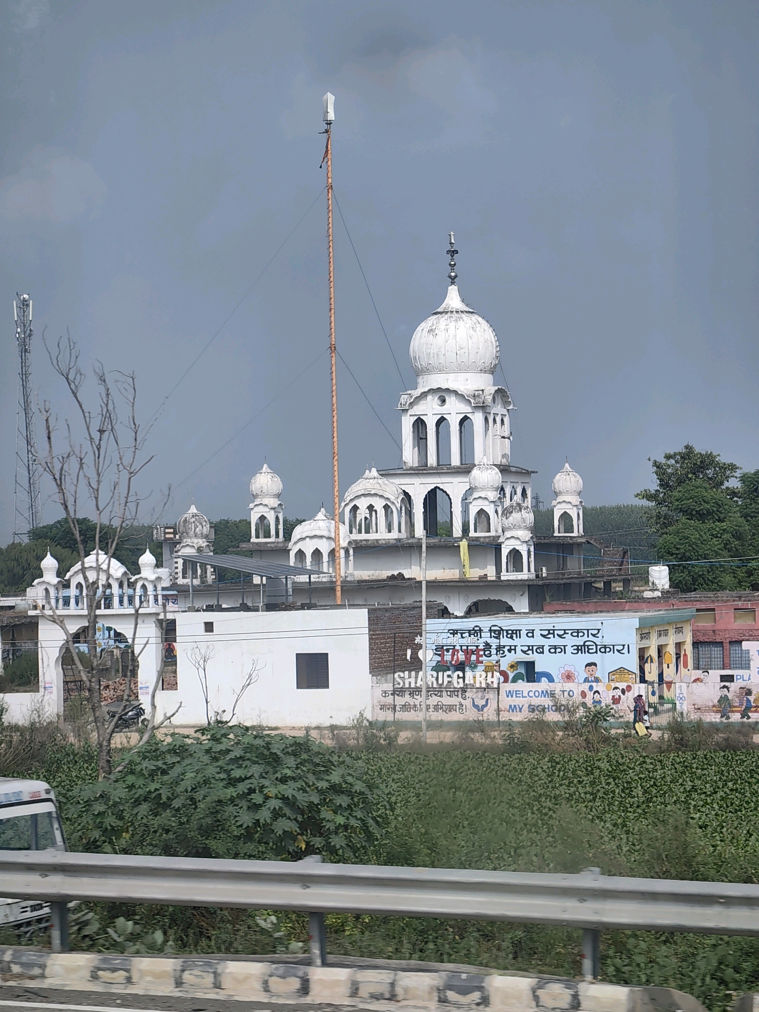 The image depicts a Gurdwara, a Sikh place of worship, in Charhuni Jattan, India. The Gurdwara is a white domed structure with multiple smaller domes and arched windows. A tall, orange pole with a flag is visible near the main dome, and a telecommunications tower stands to the left of the building. In front of the Gurdwara, there is a colorful mural on a wall, depicting children and text in Hindi and English, including the words "WELCOME TO MY SCHOOL" and "SHARIEGARH". The scene is bordered by a highway in the foreground, with dense green vegetation in the middle ground. The sky is overcast with a grey hue. There are no visible people clearly engaged in any specific actions, though some figures are present in the mural.