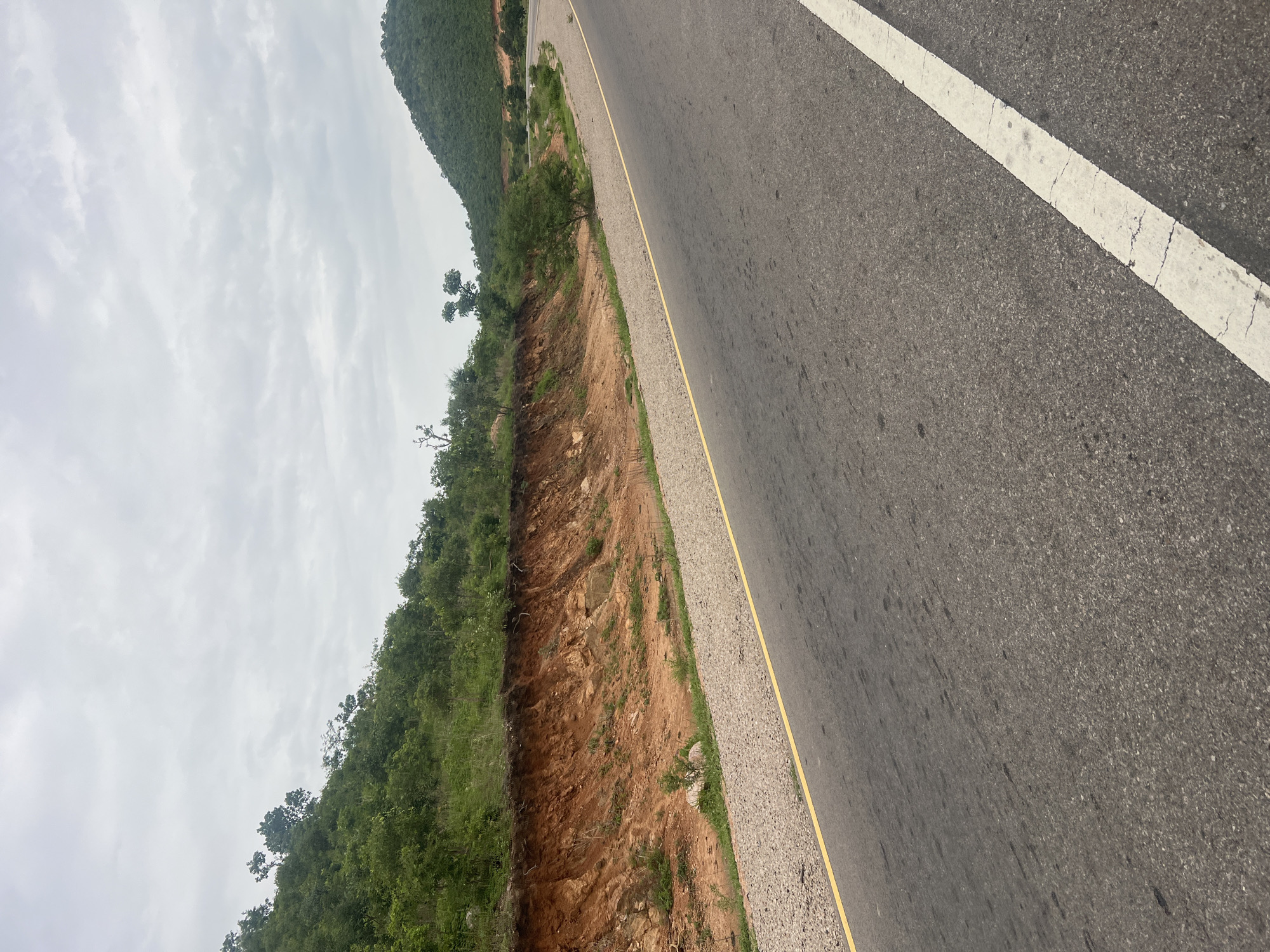 This is an eye-level, vertical, medium shot of a paved road curving through a rural landscape in Minda, Nigeria. The road is gray asphalt and has a yellow line on the left and a white line on the right. To the left of the road, a steep embankment is visible with reddish-brown soil and sparse vegetation. Above the embankment, lush green trees and bushes cover a hillside. To the right of the road, more green hills rise in the distance, with some trees scattered on their slopes. The sky is overcast with a pale, cloudy, gray hue. There are no people or vehicles visible in the image. The environment appears natural and undeveloped, suggesting a remote or less populated area.